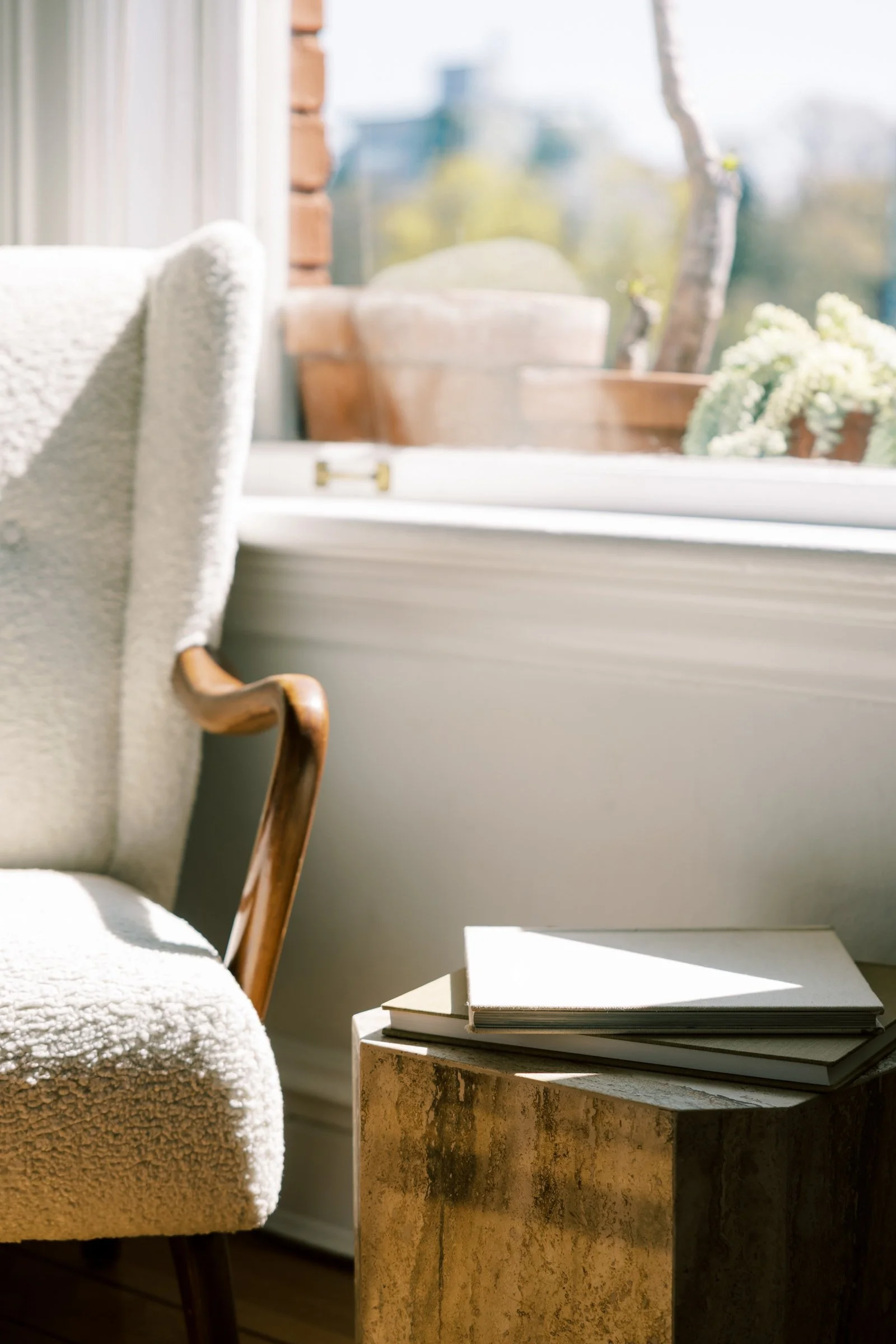 Sunlit cozy reading nook with a plush white armchair with wooden armrests and a small table with closed books, near a window showing outdoor potted plants and trees.