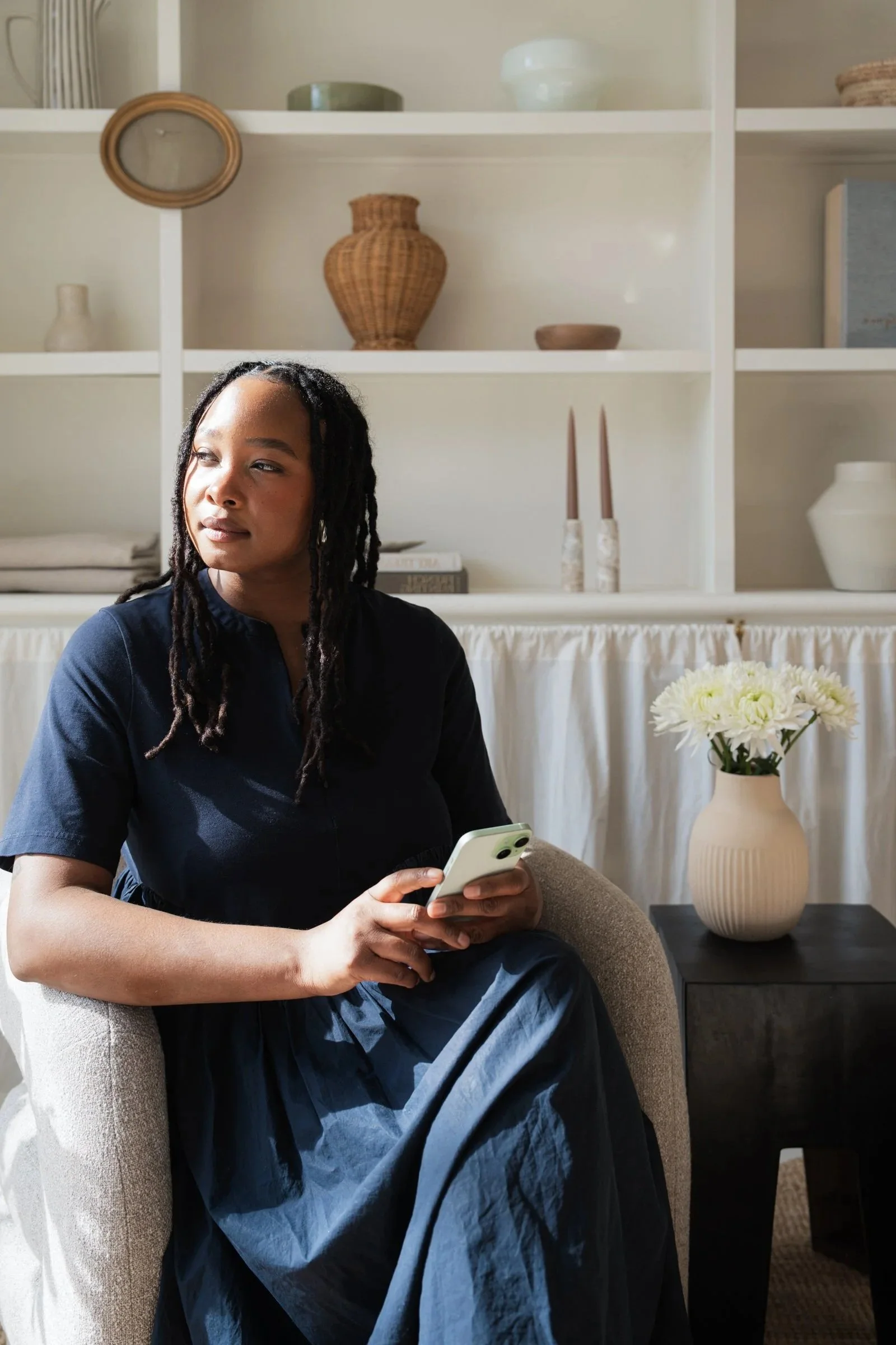 A woman with dreadlocks sitting in a beige armchair, holding a smartphone, in a living room with white shelves and decorative items in the background, including a vase with white flowers.