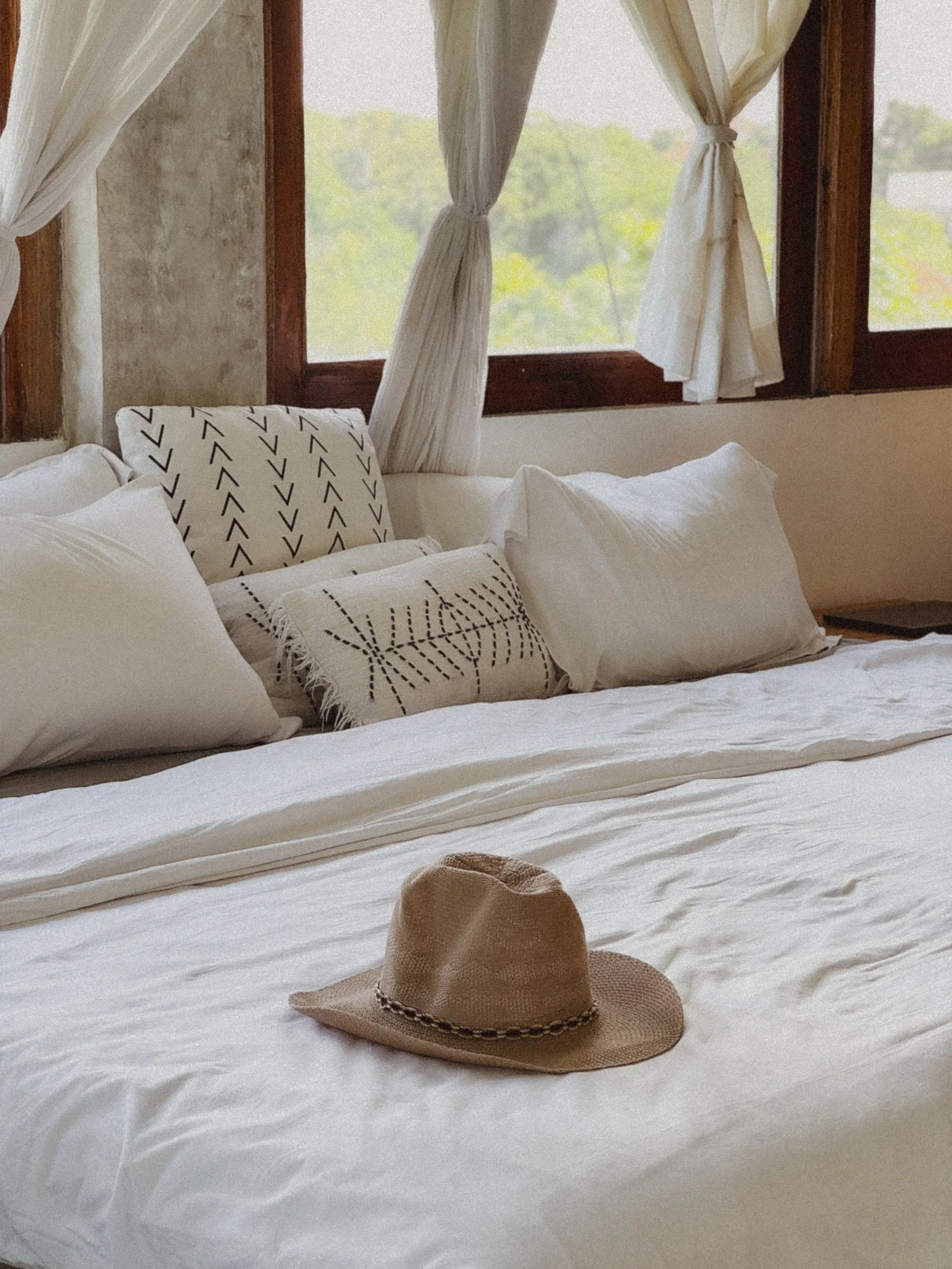 A cozy bedroom with white bed linens and various pillows, a straw hat on the bed, and windows with cream-colored curtains allowing natural light to enter.