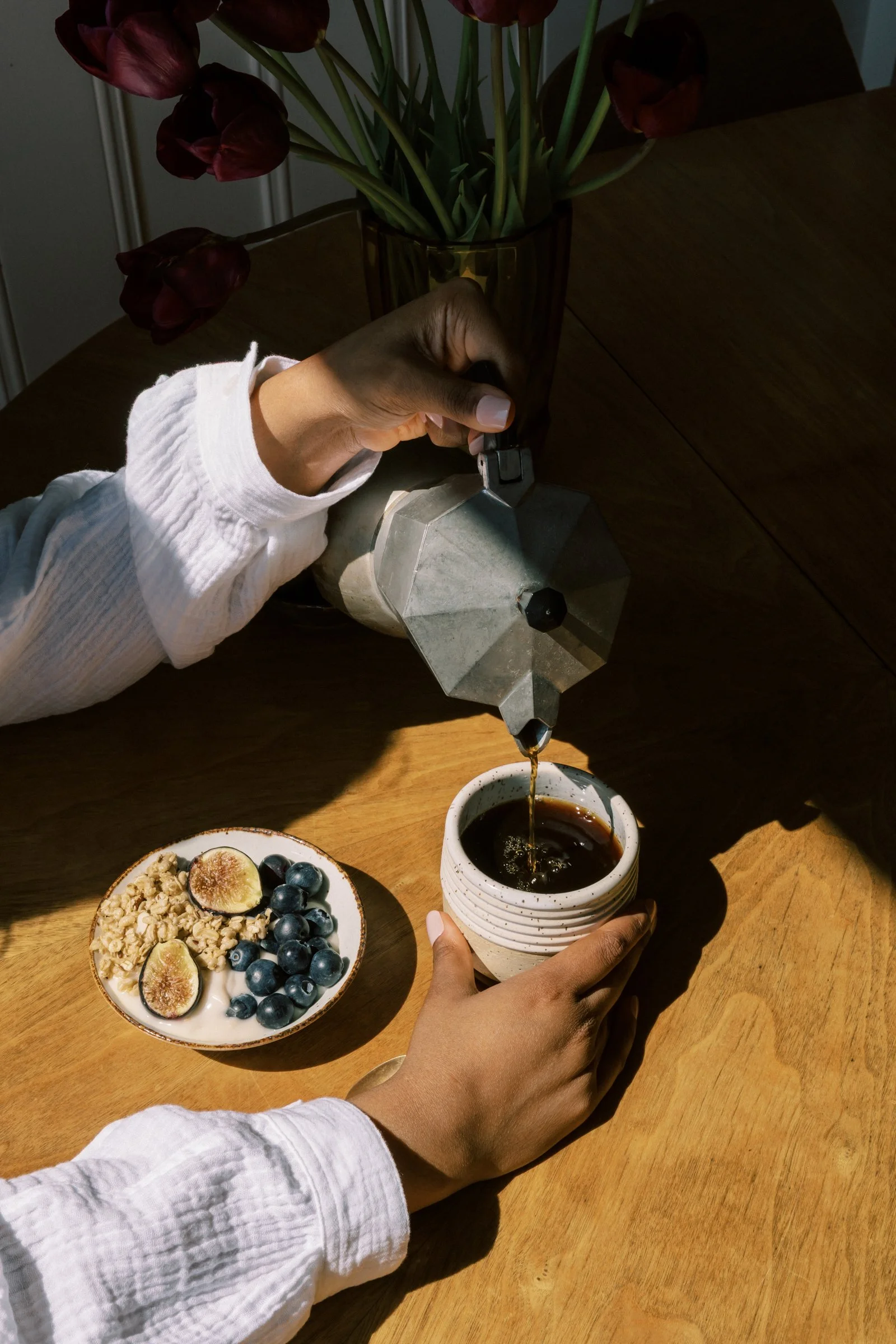 Person pouring coffee from a moka pot into a cup on a wooden table, with a bowl of yogurt topped with figs and blueberries, and a vase with dark red tulips in the background.