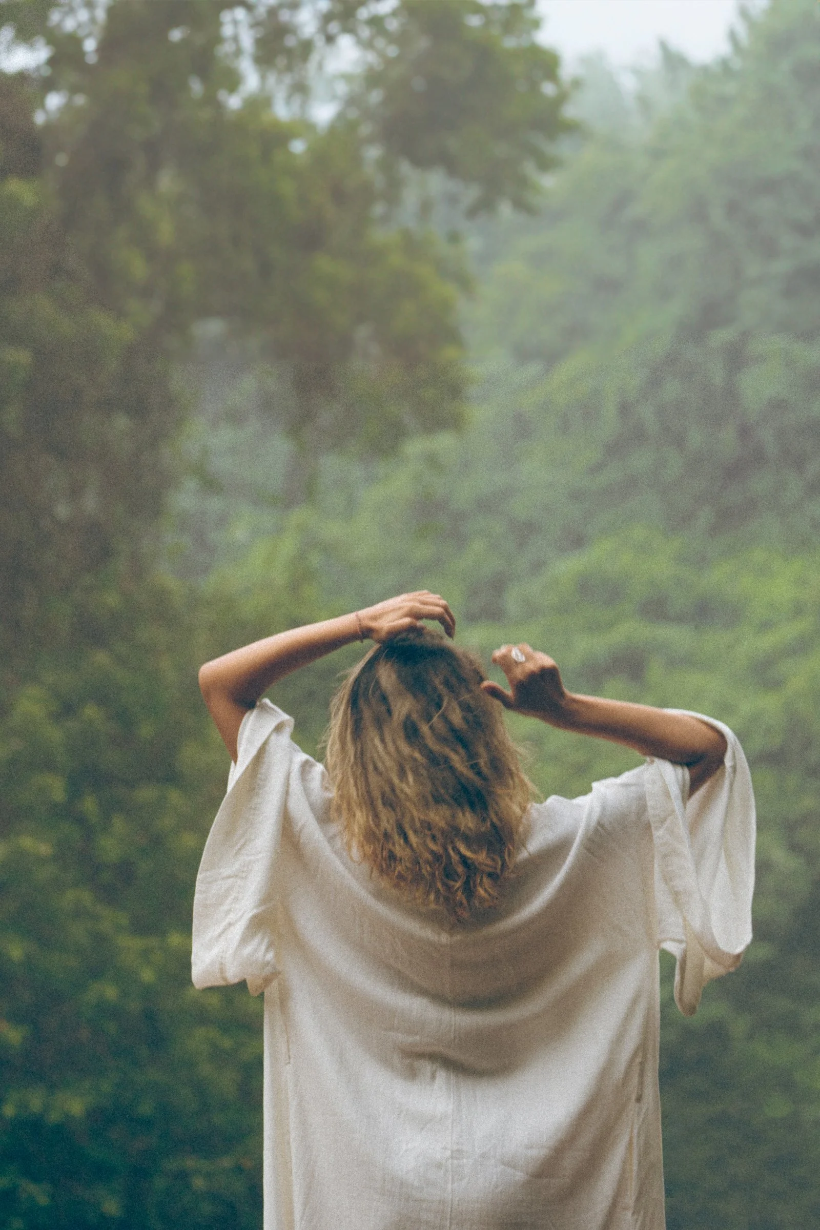 A person with curly hair wearing a white shirt, standing in a misty forest with dense green foliage, seen from behind with arms raised and hands touching their head.