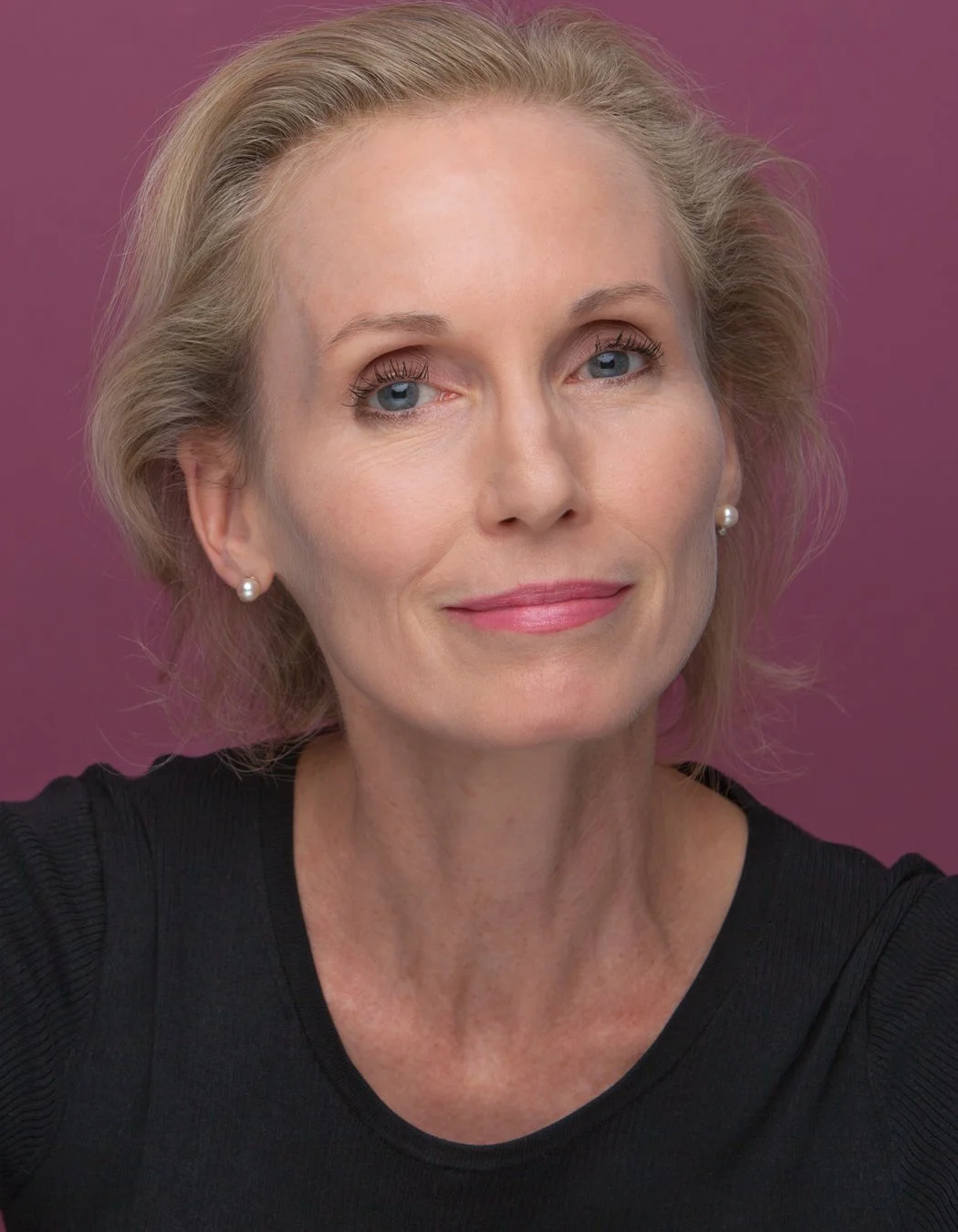 A portrait of a middle-aged woman with short blonde hair, blue eyes, and pearl earrings, wearing a black top, against a purple background.