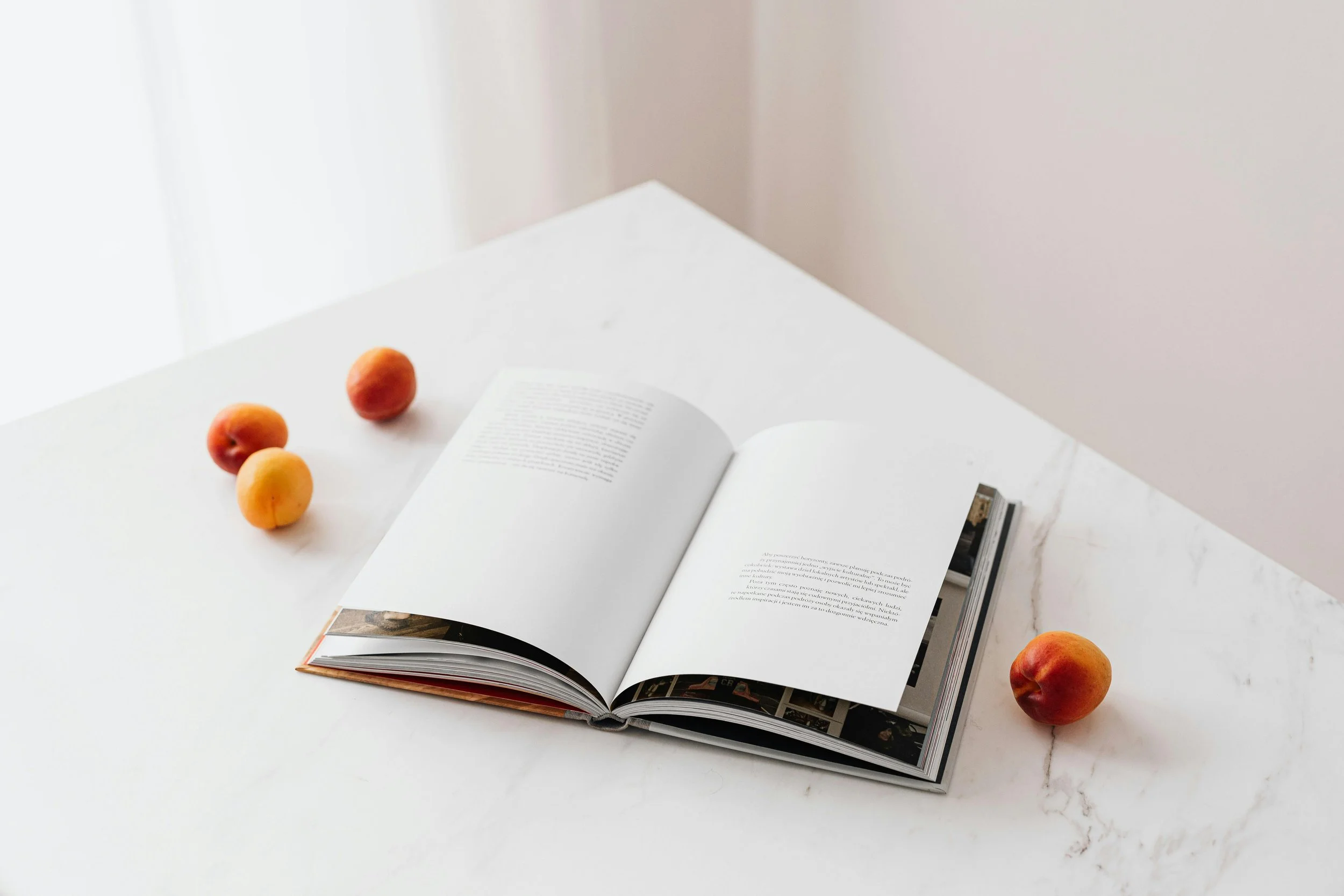 Open book on a white marble table with four peaches nearby, two on the left side and one on the right side of the table.