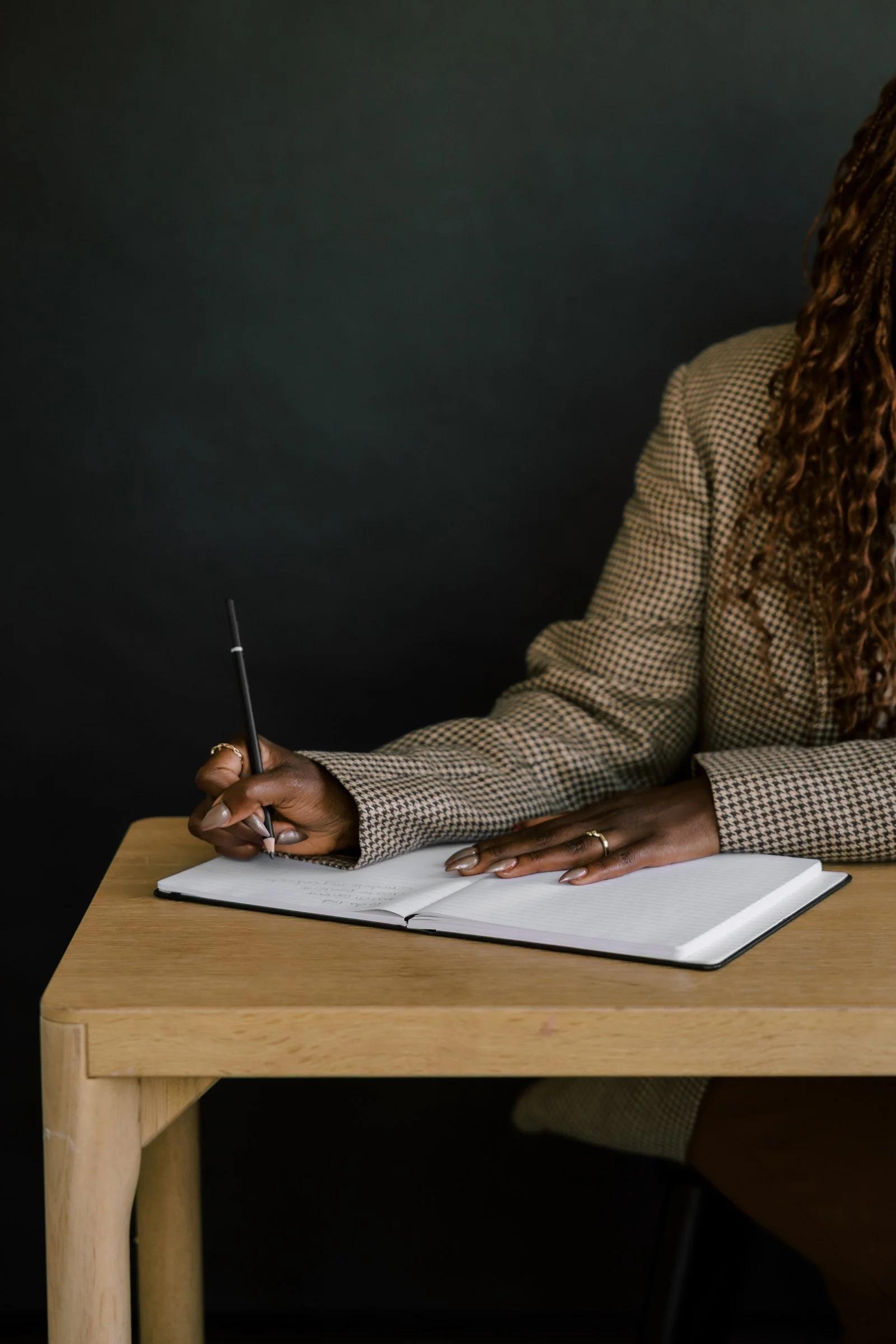 A person with curly hair in a checkered blazer sitting at a wooden desk, writing in an open notebook with a pen.
