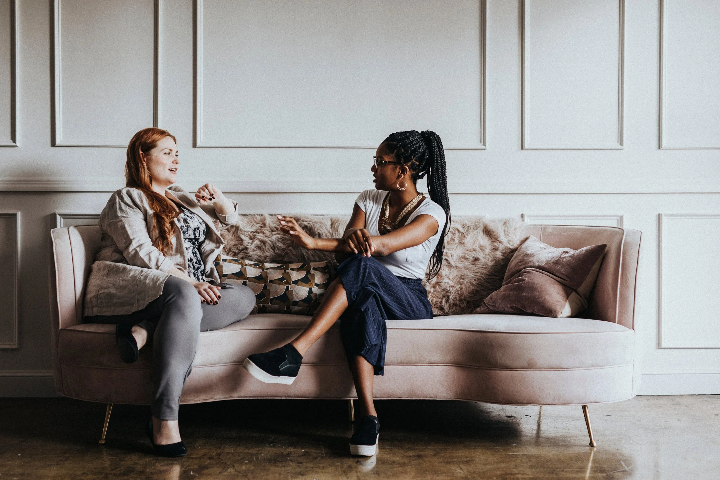 Two women having a conversation on a beige sofa in a room with white paneled walls.
