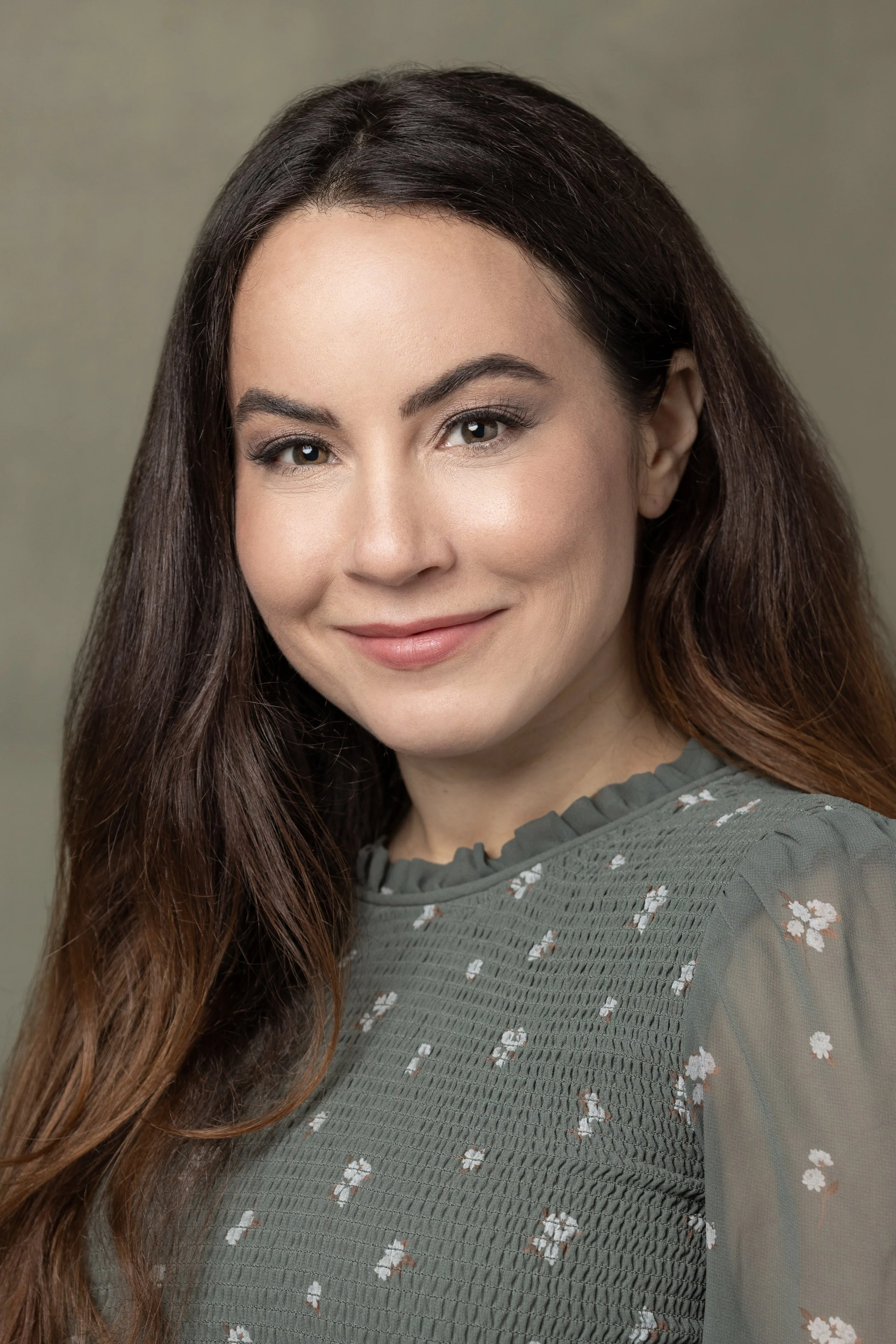 A woman with long brown hair and fair skin, smiling softly, wearing a green blouse with small white floral patterns.