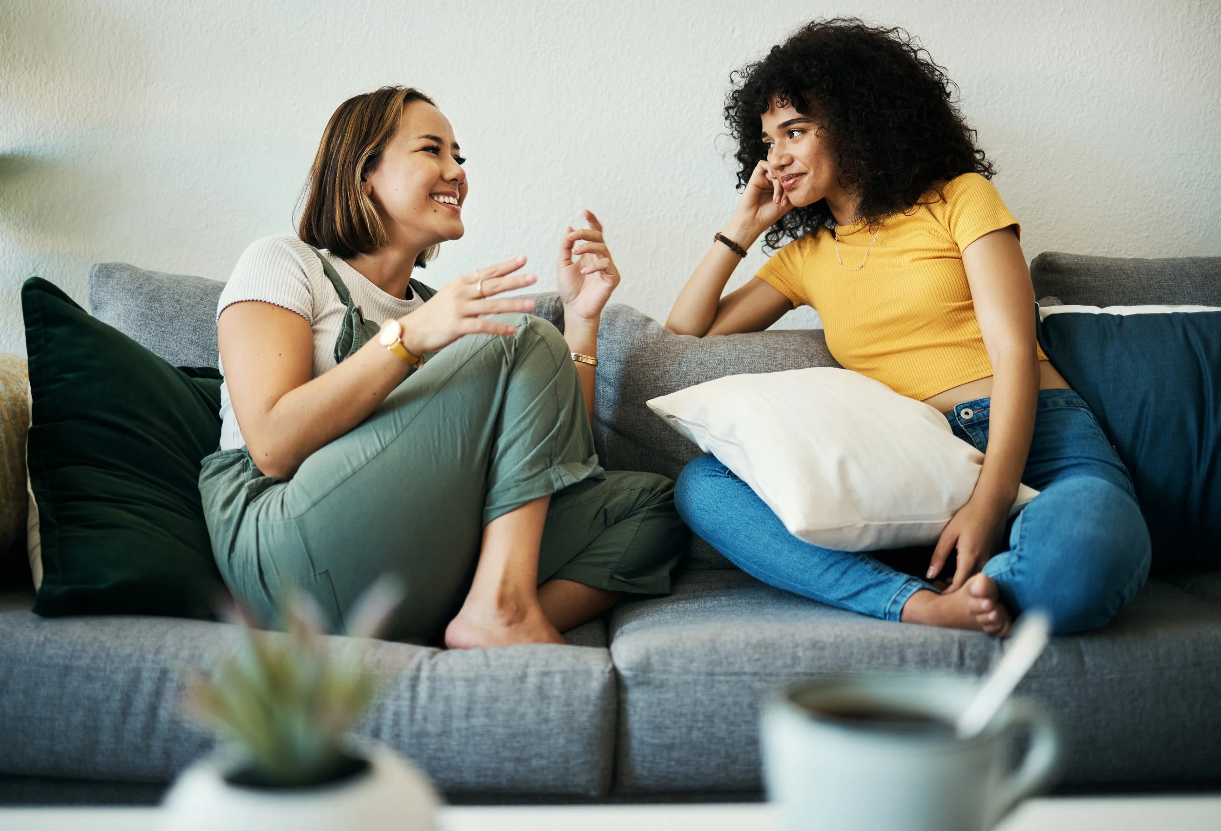 Two women conversing and smiling while sitting on a gray couch with pillows in a cozy living room.