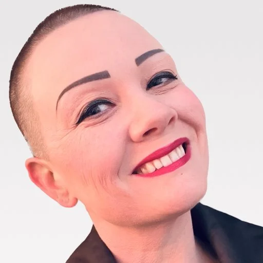 Close-up of a woman with a shaved head, wearing makeup and a black top, against a white background.