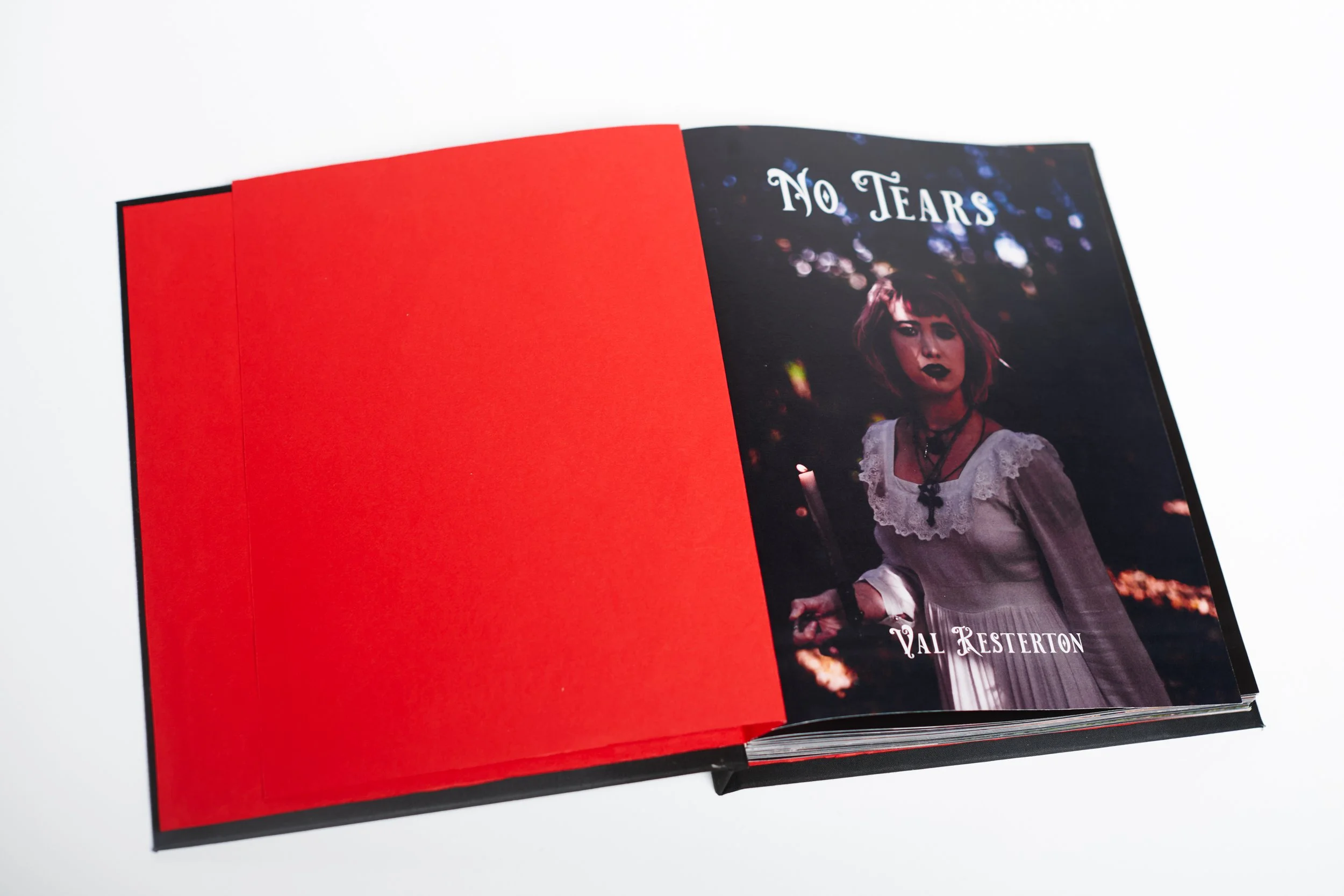 Open book with a red left page and a dark right page showing a woman wearing a white dress and black lipstick, with the text 'No Tears' and 'Val Resterton' on the right page.