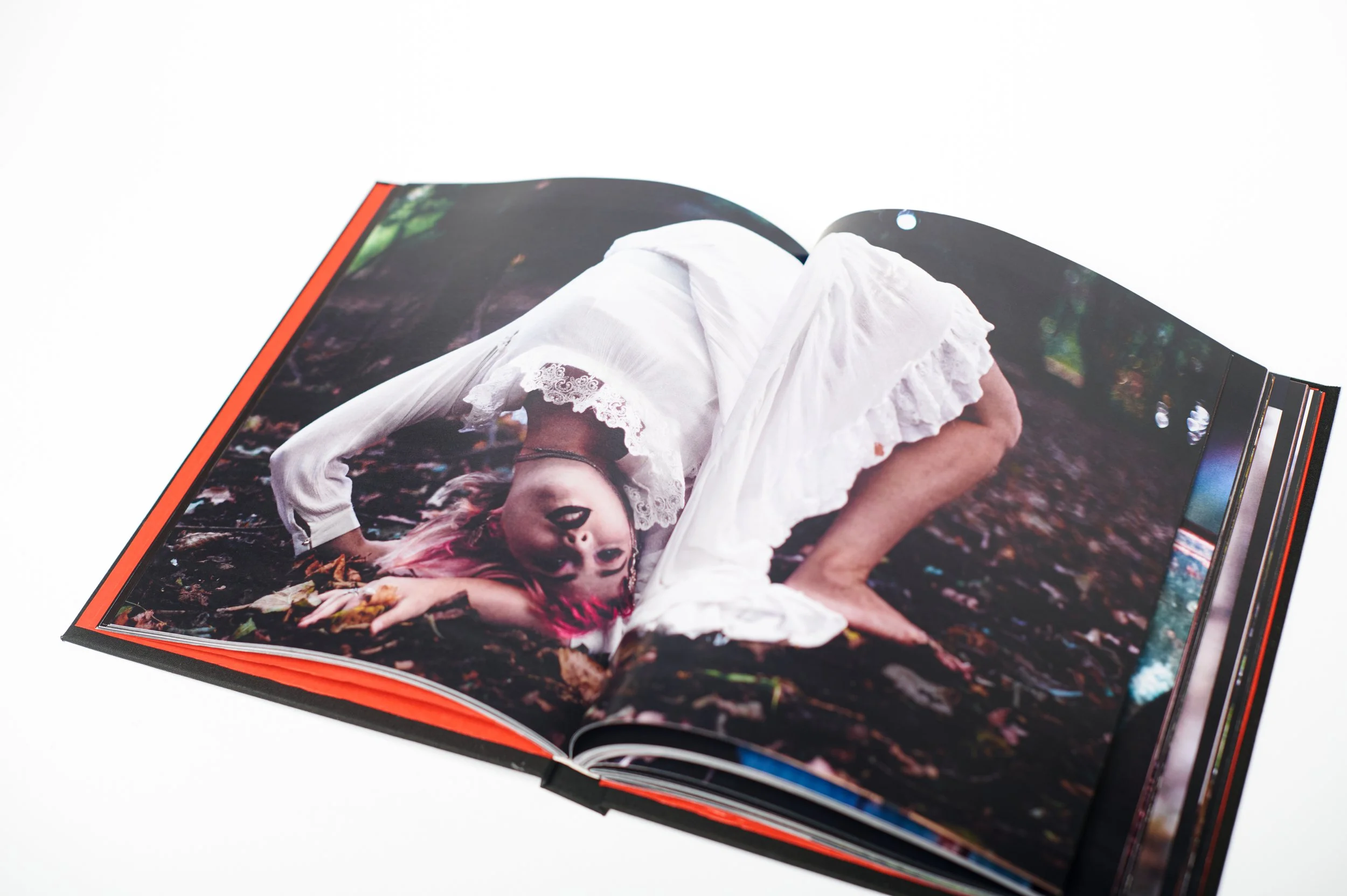 Open photo book showing a woman with pink hair in a white lace dress, lying on the forest ground surrounded by leaves.