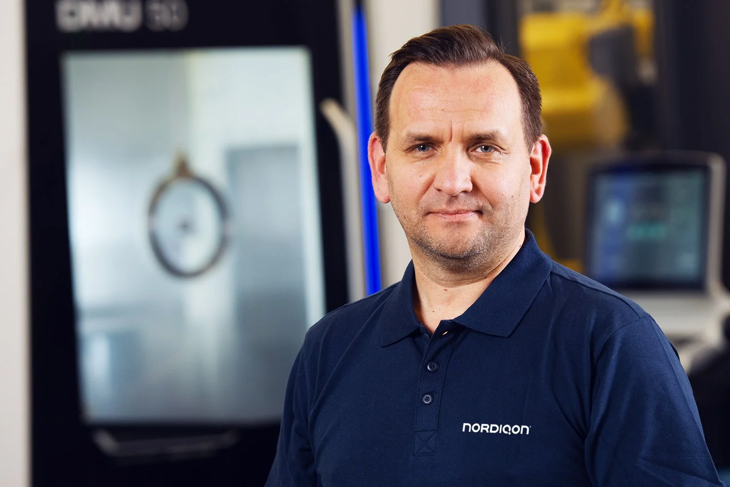 Man in navy blue polo shirt with 'Nordiqon' logo, standing in an industrial or technical environment with machinery and screens in the background.