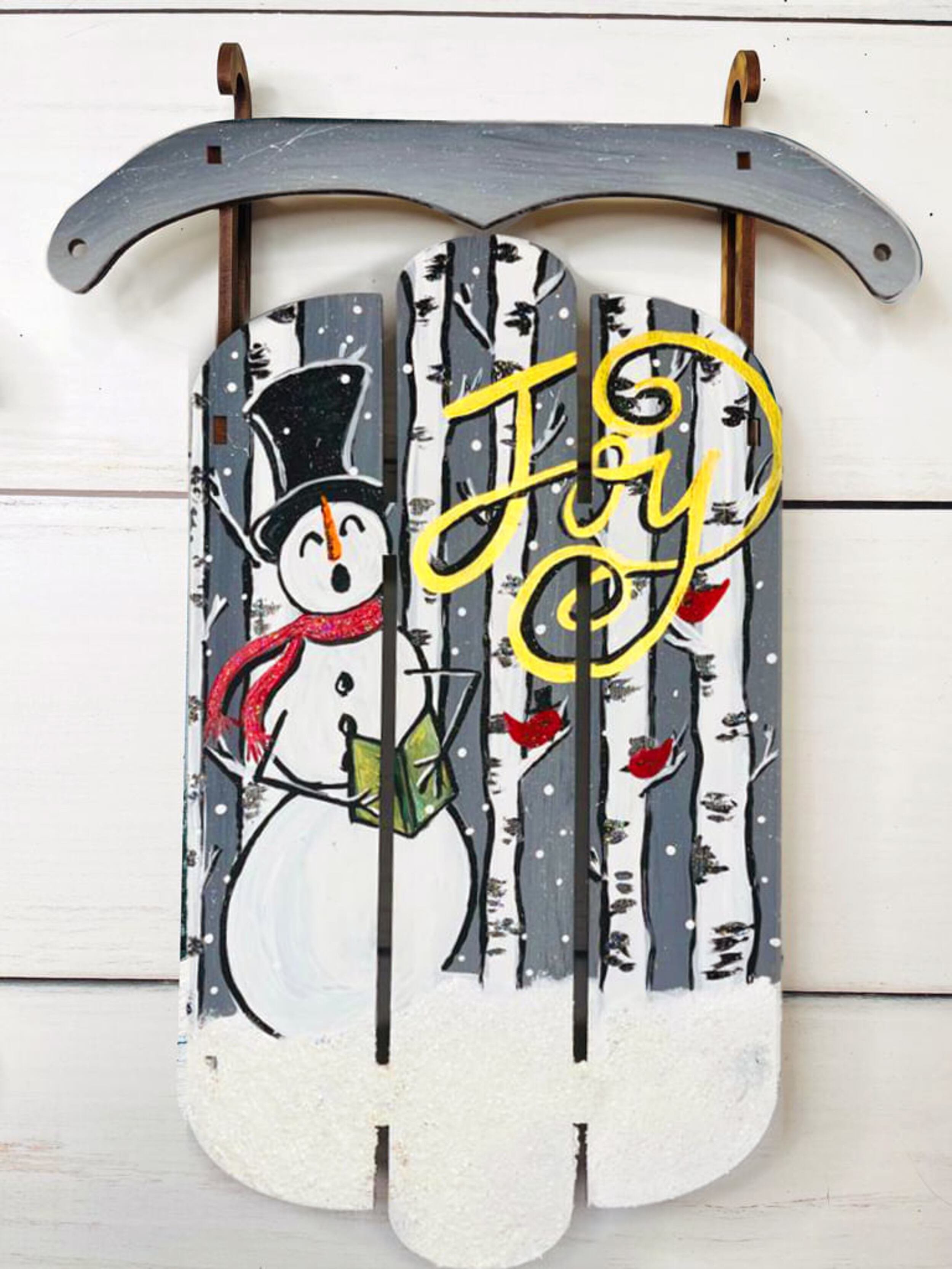 Decorative wooden sign with painted snowman wearing a black top hat and red scarf, surrounded by white birch trees with red berries in a snowy winter scene, and the words 'Joy' in yellow cursive.