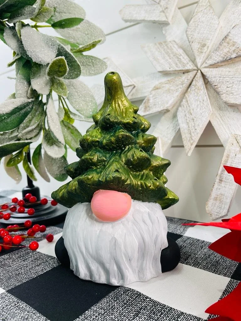 Decorative Christmas gnome with a green tree-shaped hat, pink nose, and white beard, placed on a black and white checkered tablecloth with holiday decorations around.