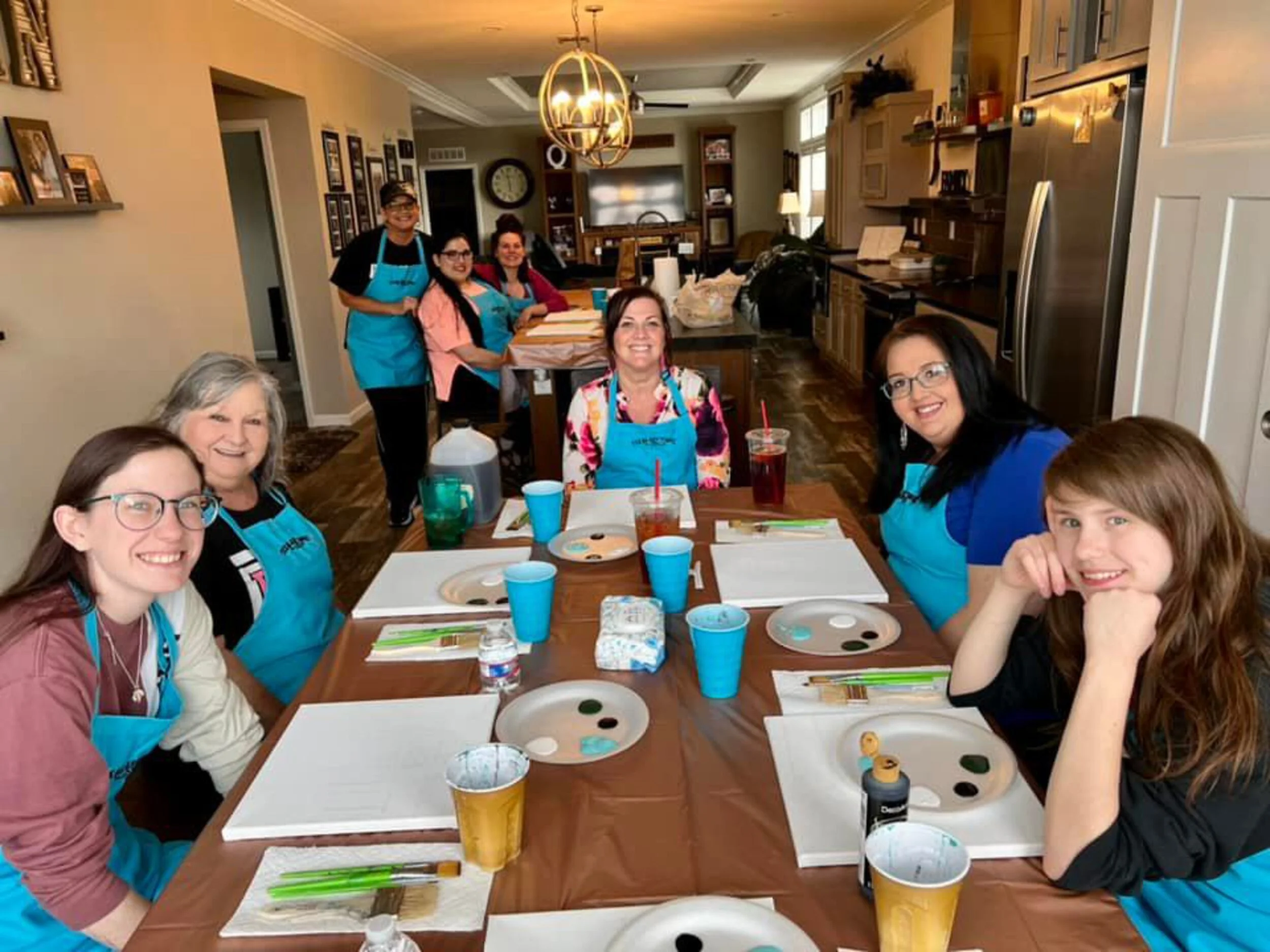 Group of women and one man sitting and standing around a dining table in a kitchen, smiling, with art supplies and drinks, during an art class or gathering.