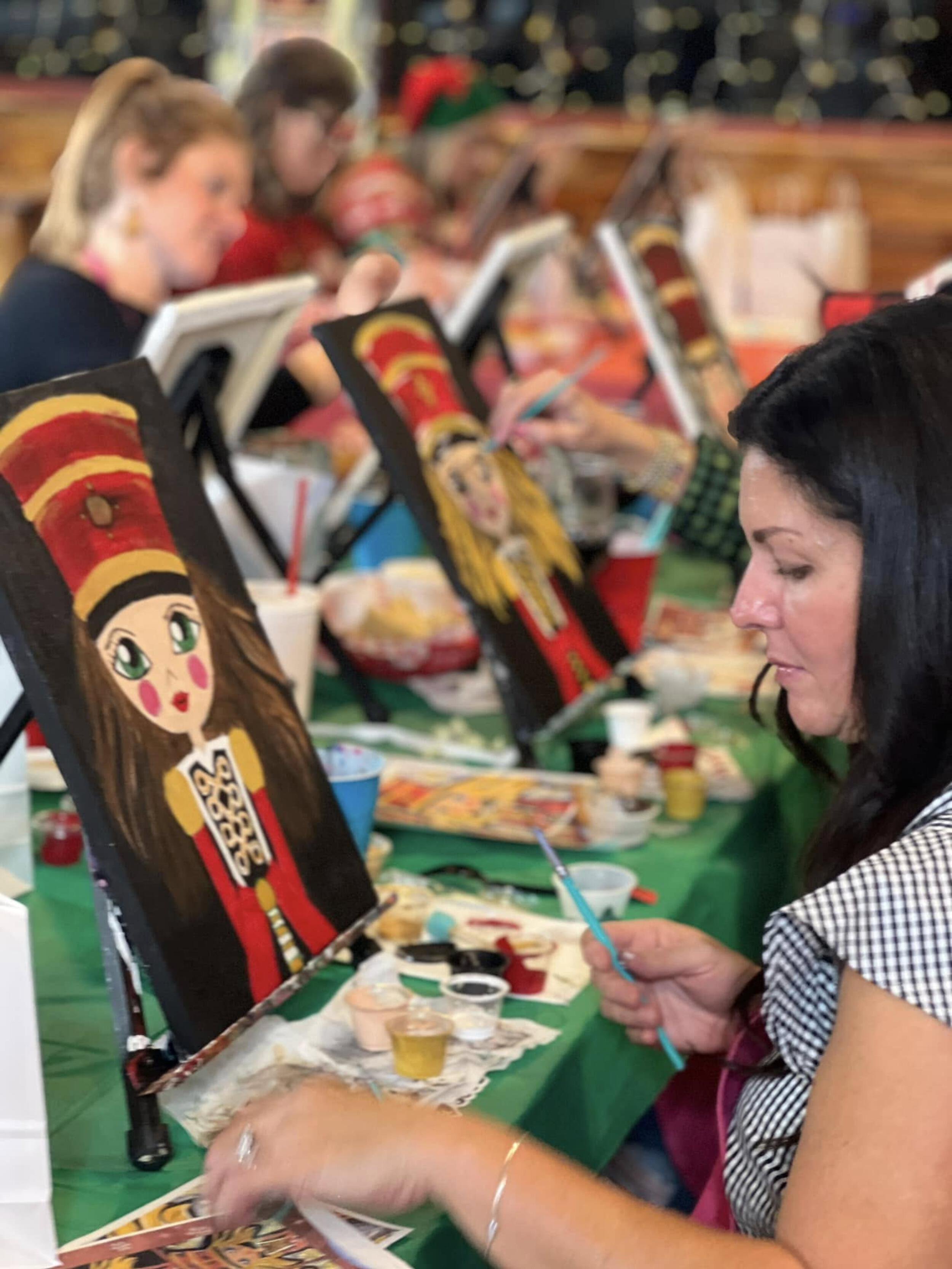 Women painting themed Christmas holiday art portraits of women in red and gold costumes on canvases at a group event.
