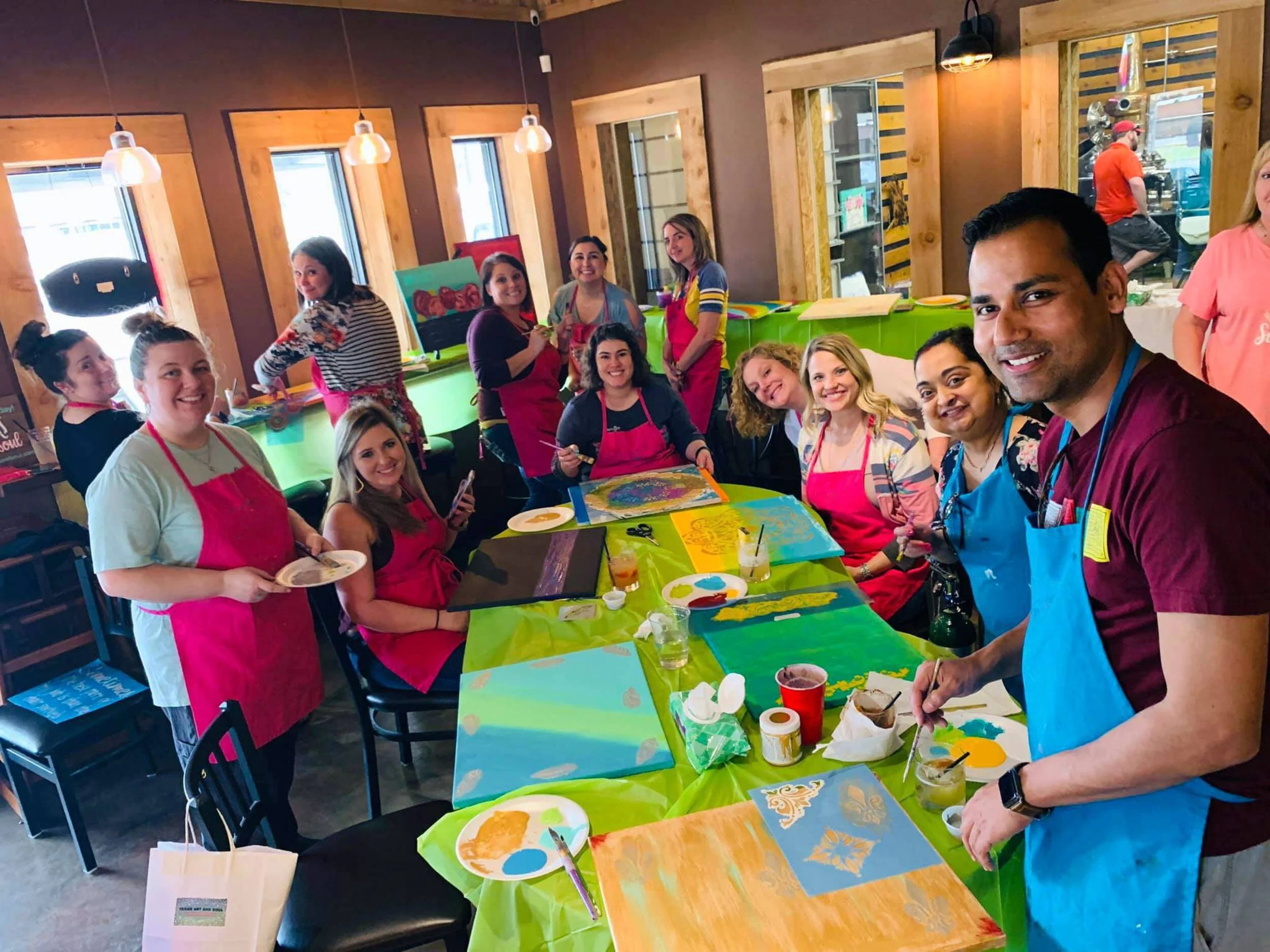 A group of people participating in a painting class, sitting and standing around a long table with colorful paintings, supplies, and drinks in a cozy, well-lit room with wooden accents.
