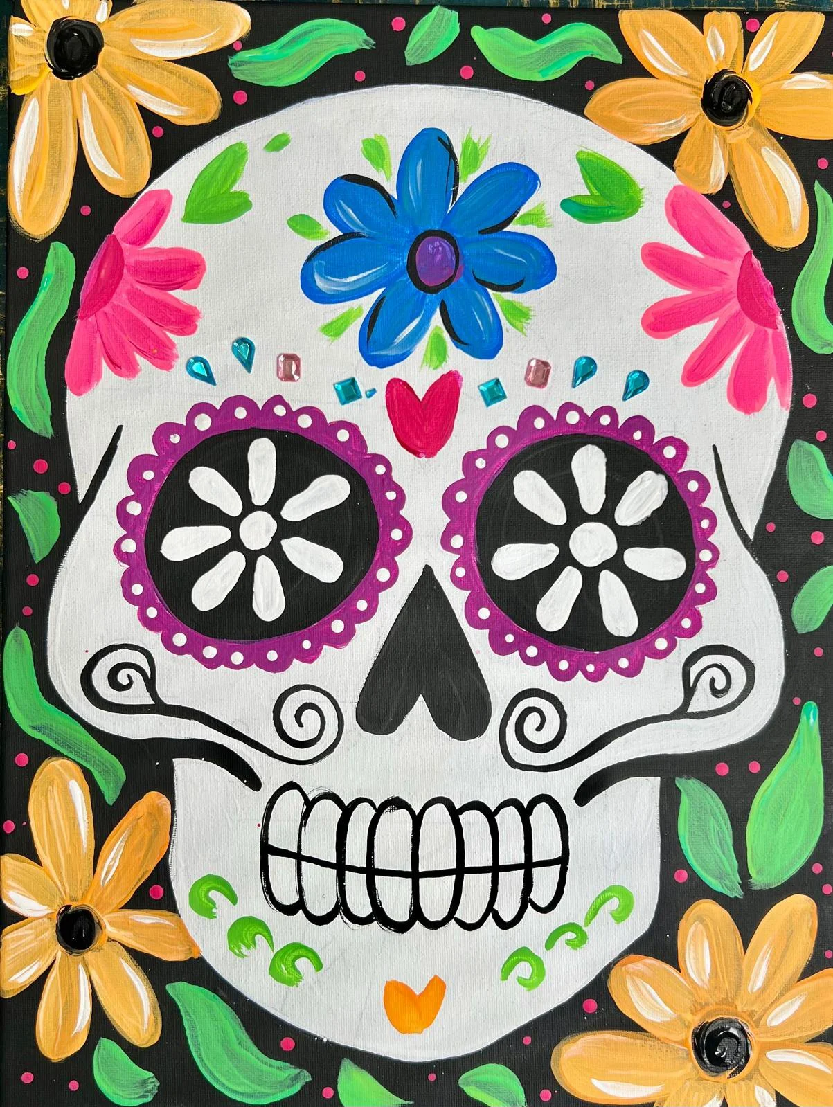 Colorful painted sugar skull with flowers and decorative designs, surrounded by orange flowers, pink accents, and green leaves on a black background, representing a Day of the Dead motif.