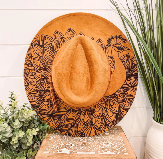 Decorative sun hat with a peacock design, surrounded by green plants, on a lace-patterned surface.