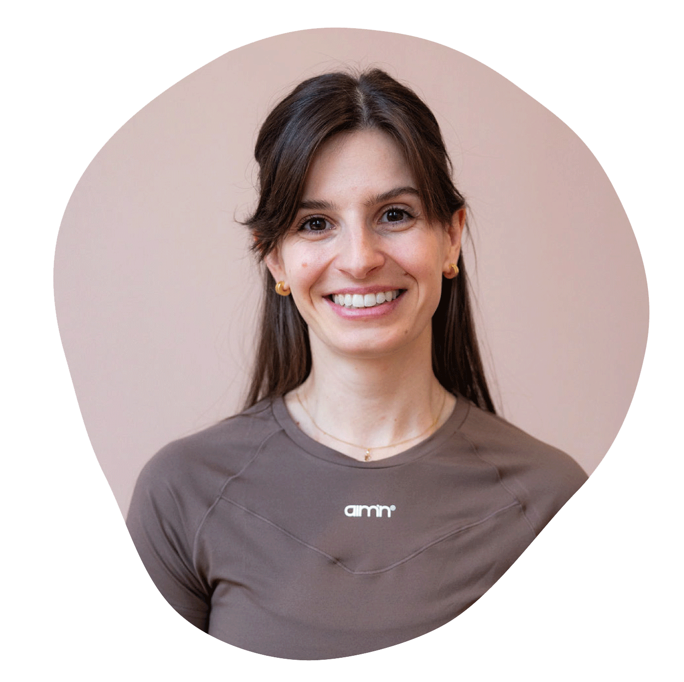 Porträt einer lachenden jungen Frau mit braunen Haaren, Ohrringen und einem hellbraunen Sport-Shirt vor einem hellen Hintergrund.