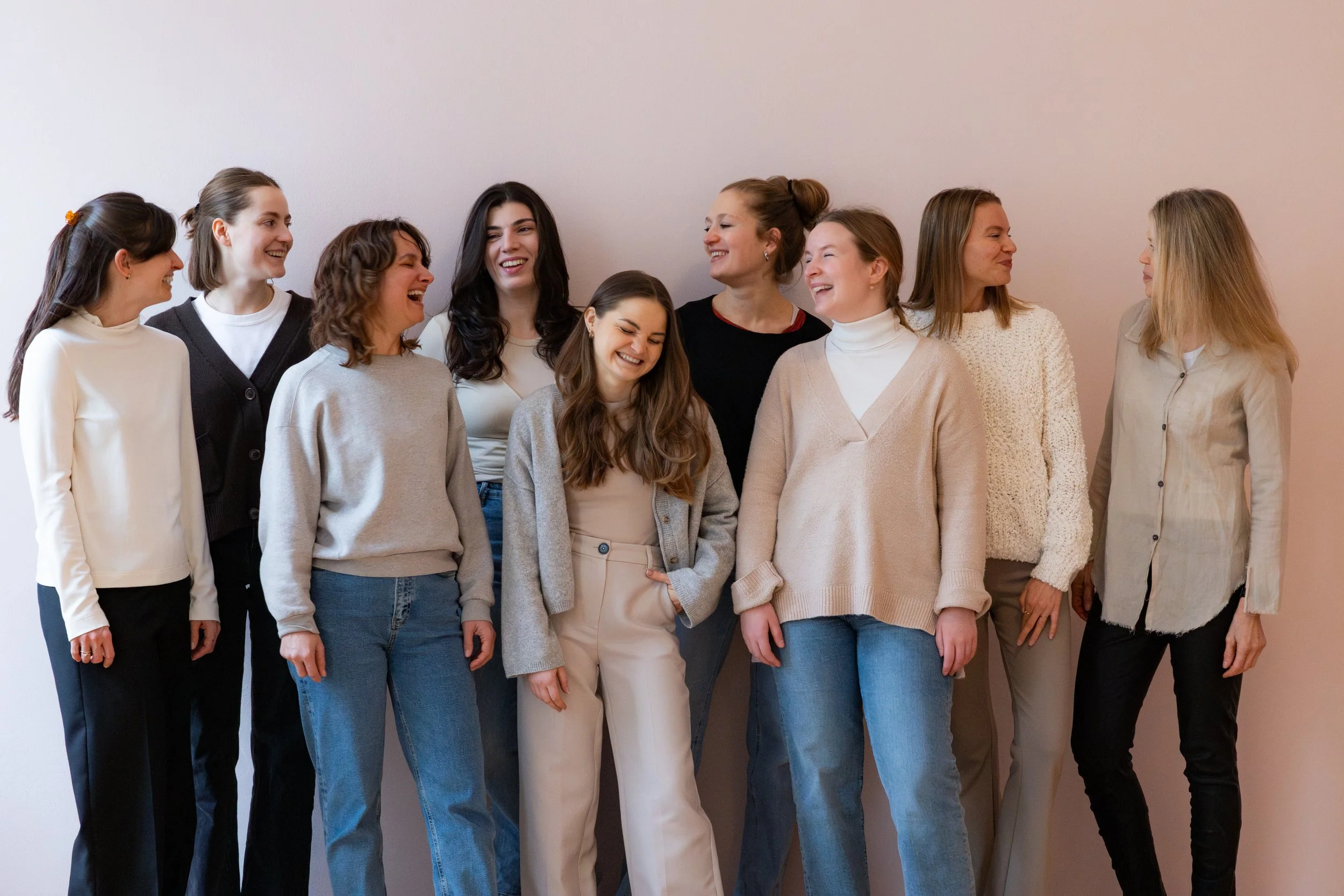Eine Gruppe von zehn lachenden Frauen steht vor einer neutralen Wand.