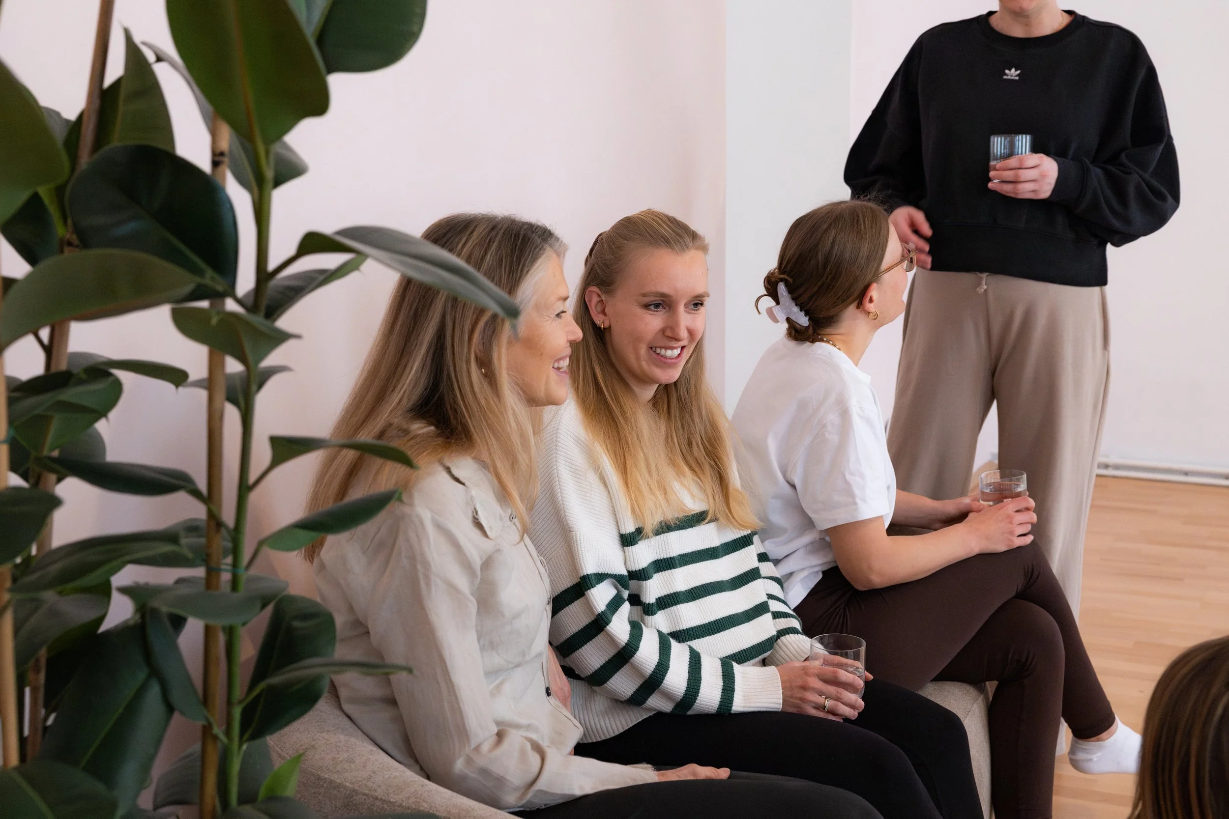 Vier Frauen sitzen auf einer Couch und eine Person steht daneben, alle halten Gläser. Eine Frau trägt eine weiße Bluse, eine trägt ein gestreiftes Hemd, eine andere eine weiße Bluse, und die vierte Person trägt ein schwarzes Sweatshirt. Es gibt eine große grüne Pflanze im Vordergrund.