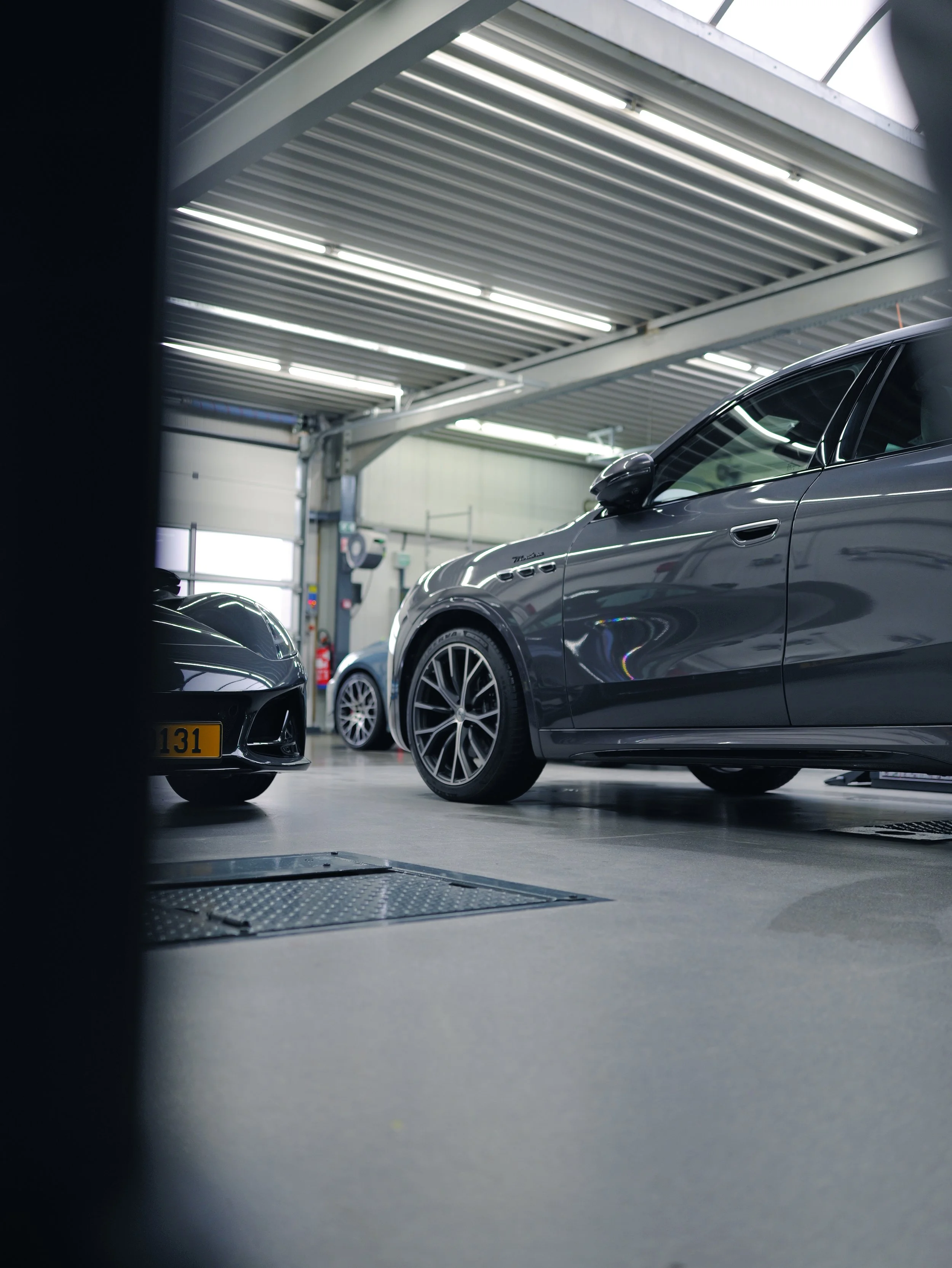 Voiture gris foncé dans un garage, vue en contre-plongée, avec un autre véhicule noir partiellement visible à gauche.