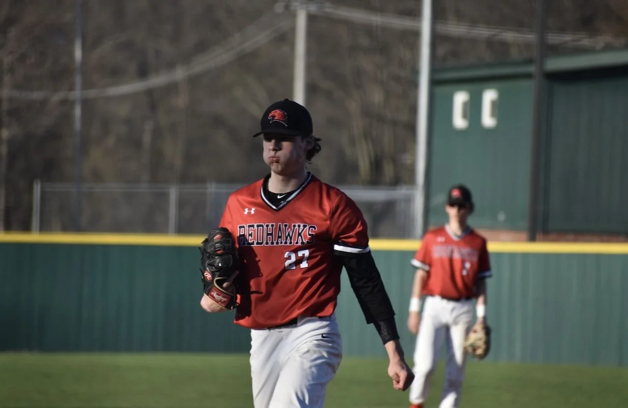 Baseball players in red and black uniforms on a field with a green fence in the background.
