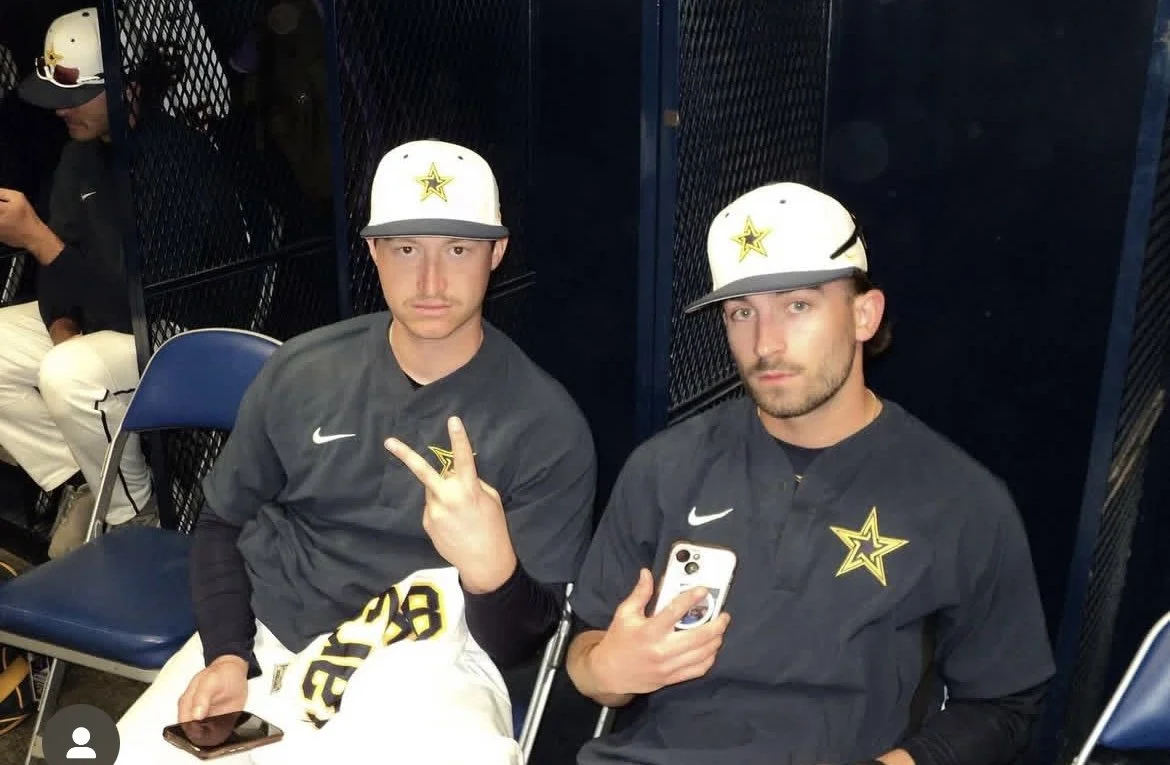 Two baseball players in black jerseys with a yellow star logo, sitting in a locker room. One makes a peace sign while the other holds a phone. Both wear white caps with the same logo.