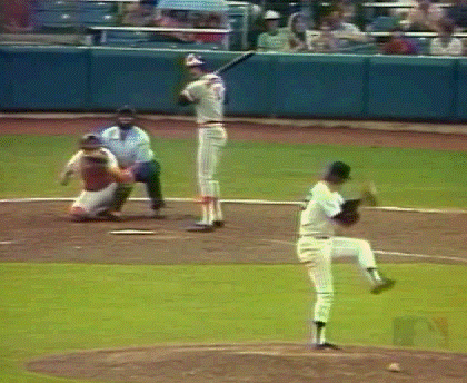 A baseball pitcher on the mound in the act of pitching, a batter preparing to swing, and an umpire behind the batter during a game.