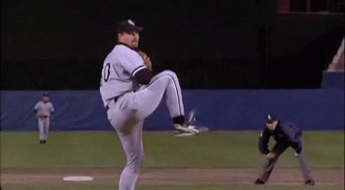 A baseball pitcher mid-throw on the mound with an umpire and other players nearby at a baseball game.