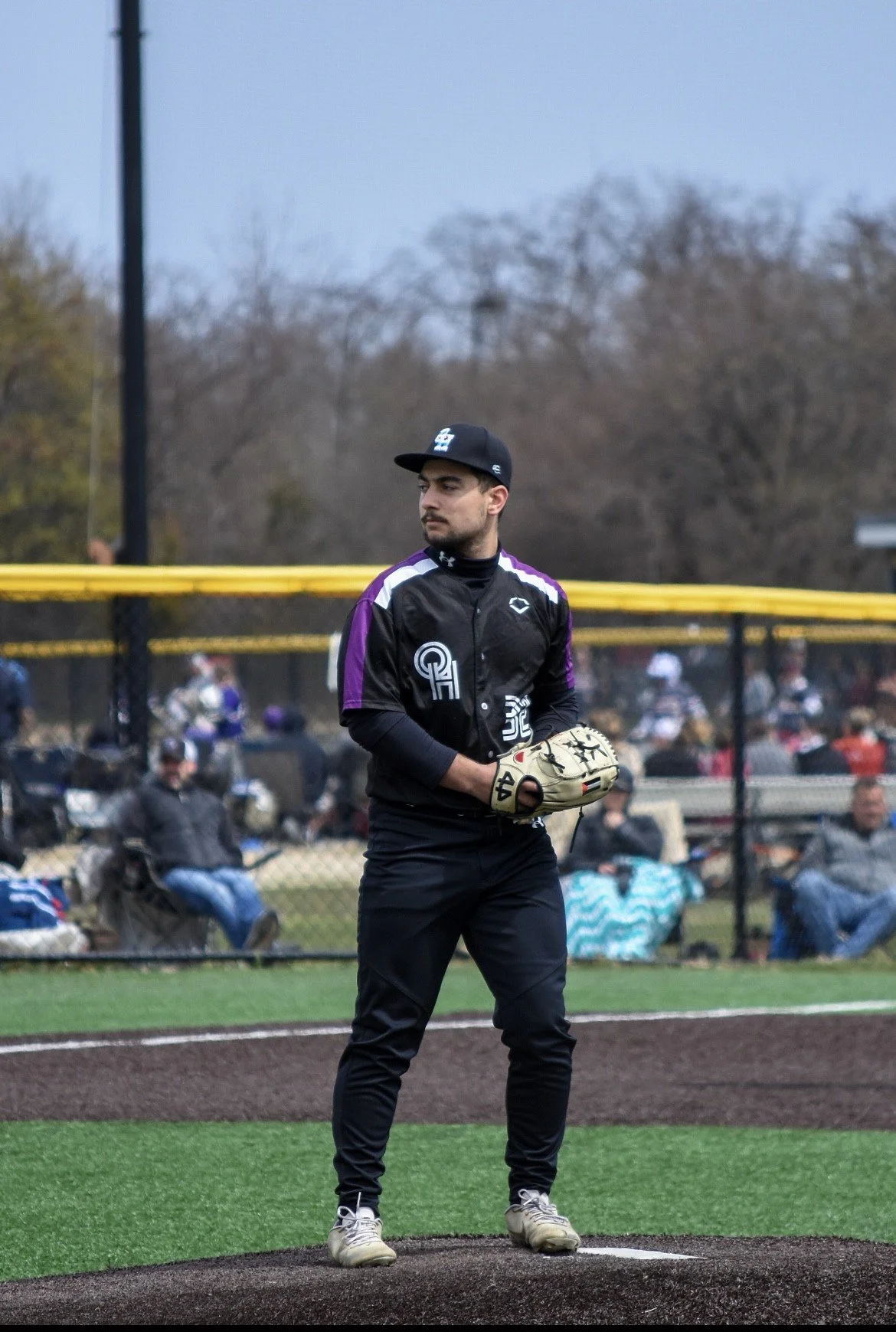 A baseball player standing on the pitcher's mound, holding a baseball glove, with a baseball field and spectators in the background.