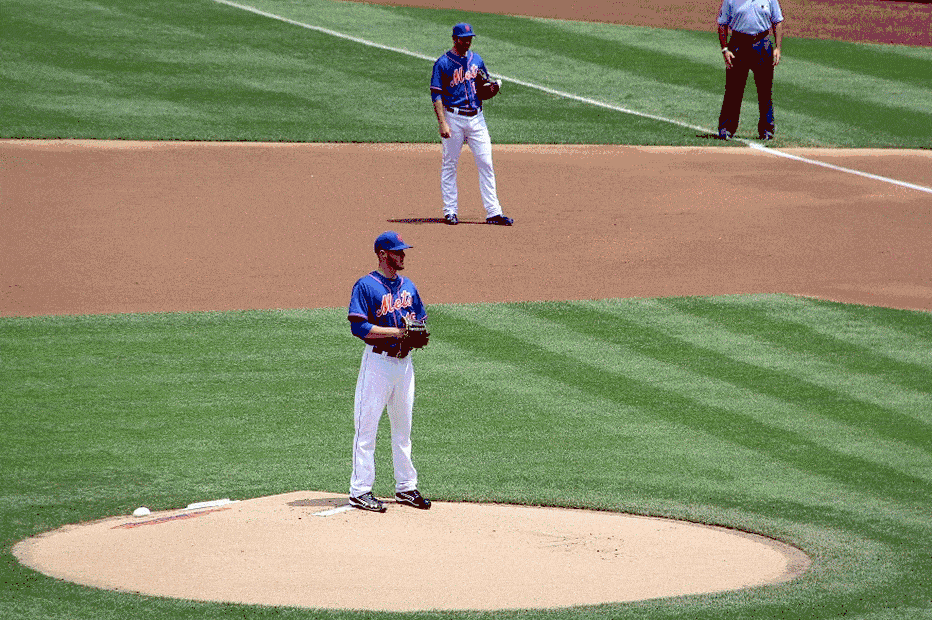 A baseball pitcher stands on the pitcher's mound with a ball in hand, preparing to pitch during a game. Another player is positioned behind him on the field, and an umpire is visible in the background.