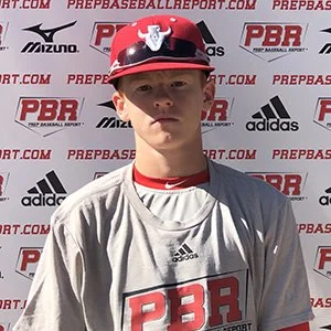 Young baseball player wearing a red and black helmet, gray jersey with PBR logo, standing in front of a PBR and Adidas branded backdrop.
