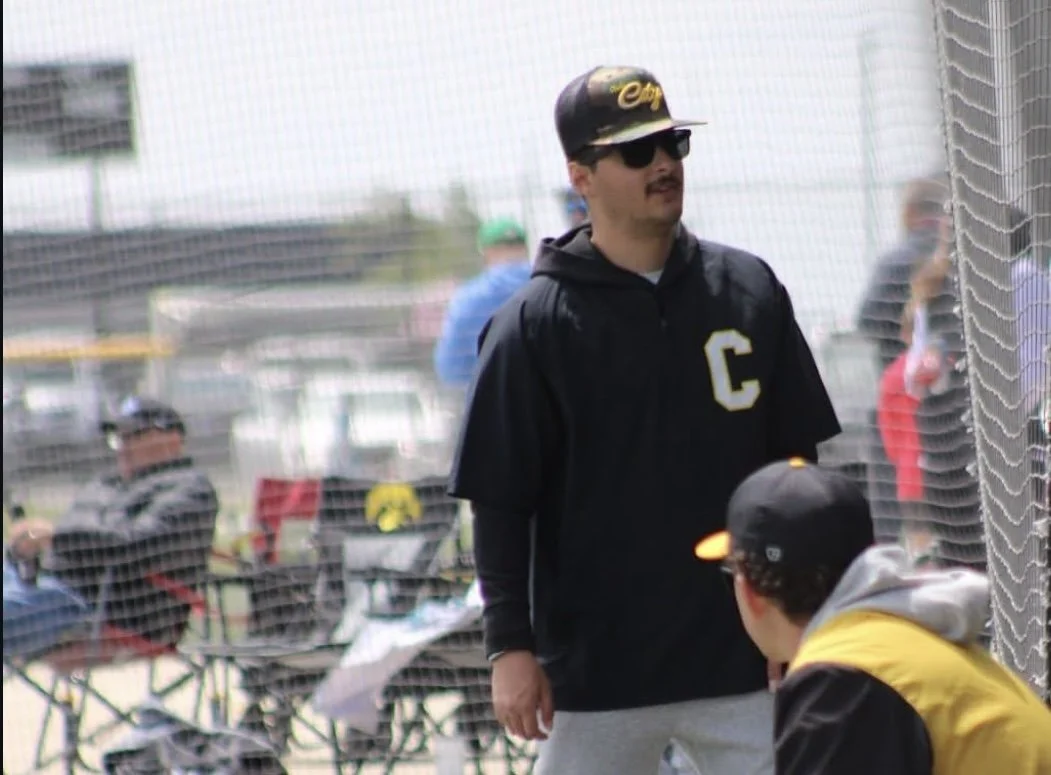 A man wearing sunglasses, a black hoodie with a C on it, black cap with gold lettering, and gray sweatpants, standing near a baseball net during a game, engaged in conversation with another person wearing a black cap, yellow and black jacket.