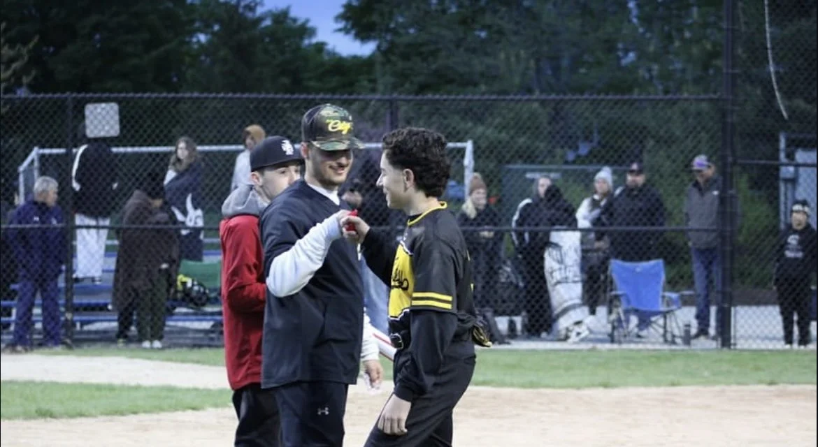 Three young men are at a baseball field, with the middle one holding a medal or award, and the one in the black and yellow jersey is smiling and shaking hands with him. Spectators and opponents are in the background behind a chain-link fence.