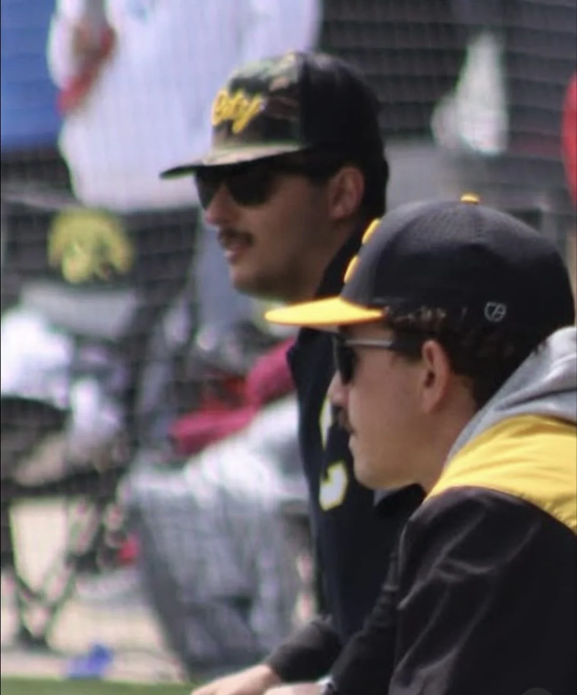 Two men sitting side by side, wearing black and yellow caps and sunglasses, outdoors with blurred people in the background.