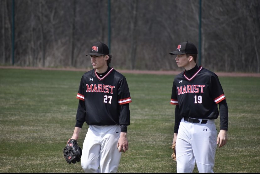 Two young baseball players in black and red jerseys, black hats, and white pants walking on a baseball field.