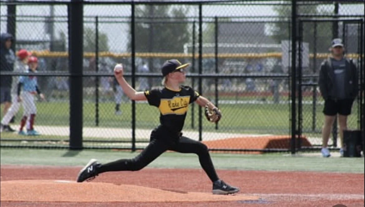 A young girl pitching a baseball on a baseball field during a game or practice. She is wearing a black and yellow uniform and cap, and is in mid-throw with her right arm extended.