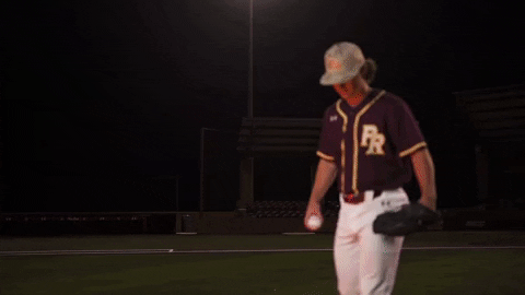 A young baseball player wearing a maroon jersey, white pants, and a hat, standing on a baseball field at night, holding a baseball in one hand and a glove in the other.