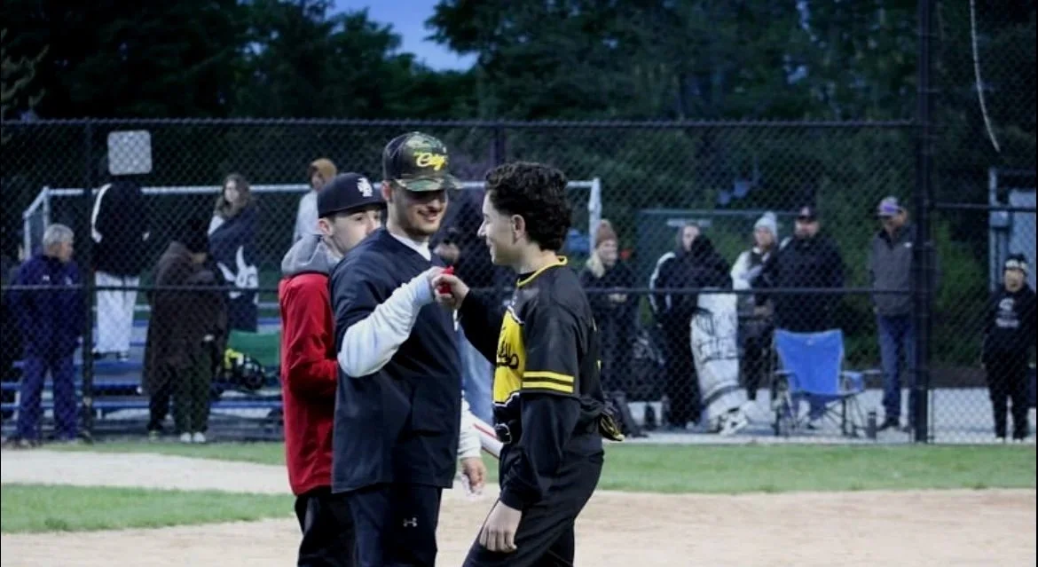 Two young men shaking hands on a baseball field, with a group of spectators behind a chain-link fence in the background.