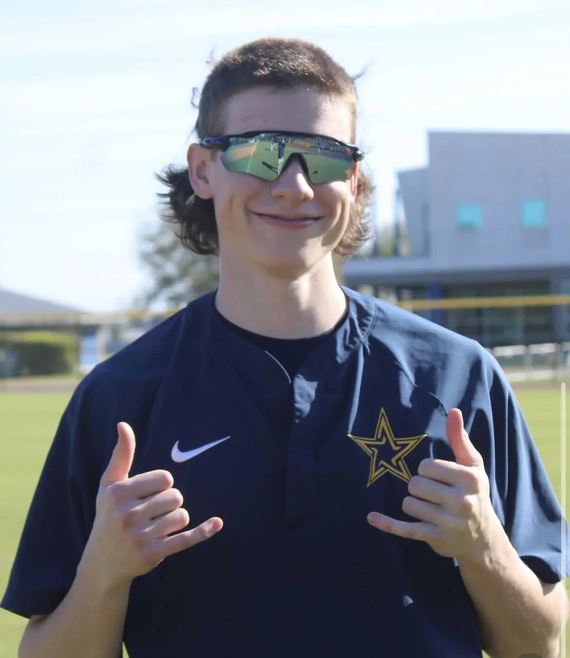 A young man wearing sports sunglasses and a navy athletic jacket with a star logo, standing outdoors on a sunny day, giving two thumbs up.