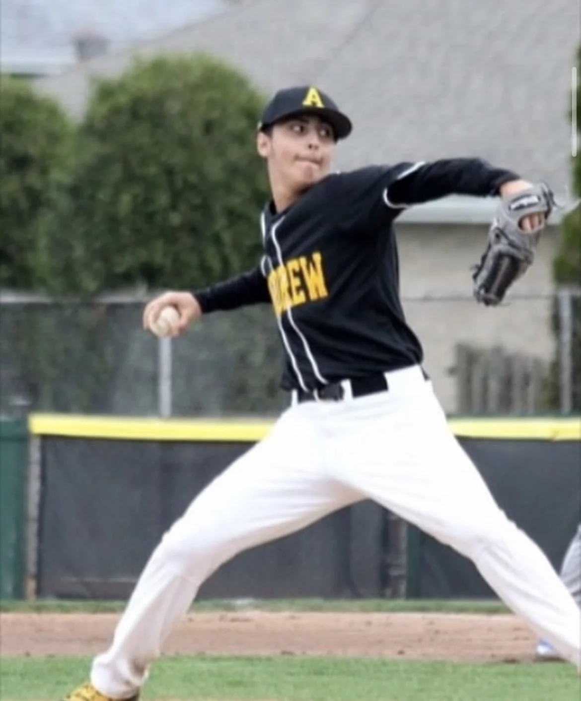 Young baseball pitcher in uniform is mid-throw on a baseball field, wearing a black cap and shirt, white pants, and holding a baseball and glove.