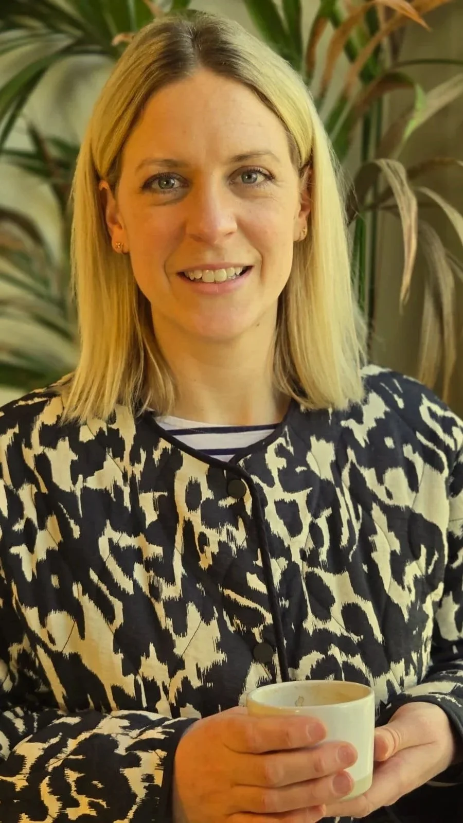 A woman with shoulder-length blonde hair, smiling and holding a white mug, standing in front of leafy green plants.