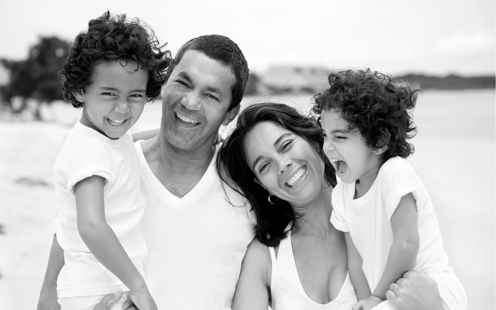 My Ideal Parents - PBSP - A black-and-white photograph of a smiling family of four enjoying time together at the beach. The two children are being carried by their parents, and all family members are wearing white clothing.