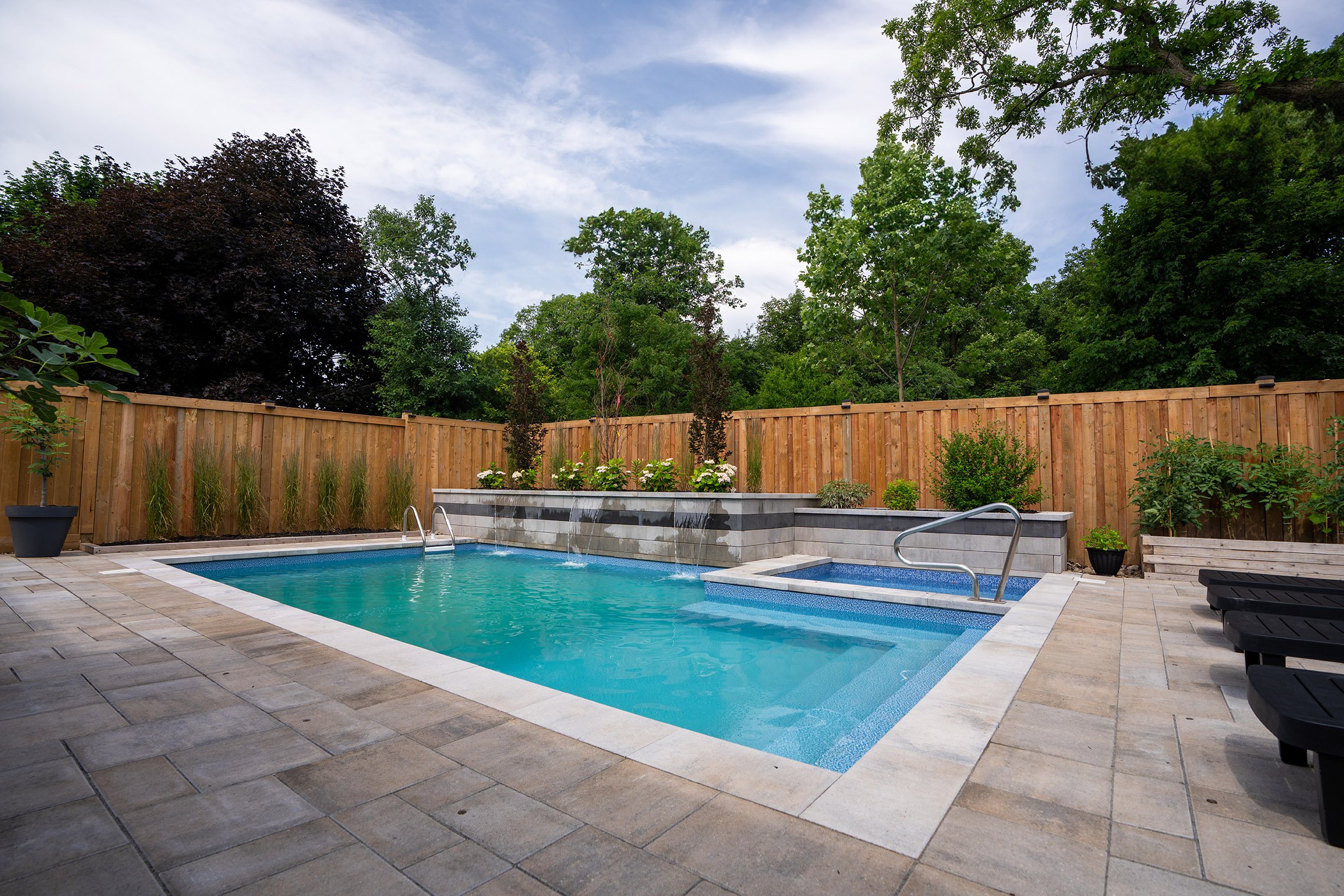 Empty swimming pool with blue water, metal ladder, concrete pool deck, wooden fence, and grassy area with some trees in the background.