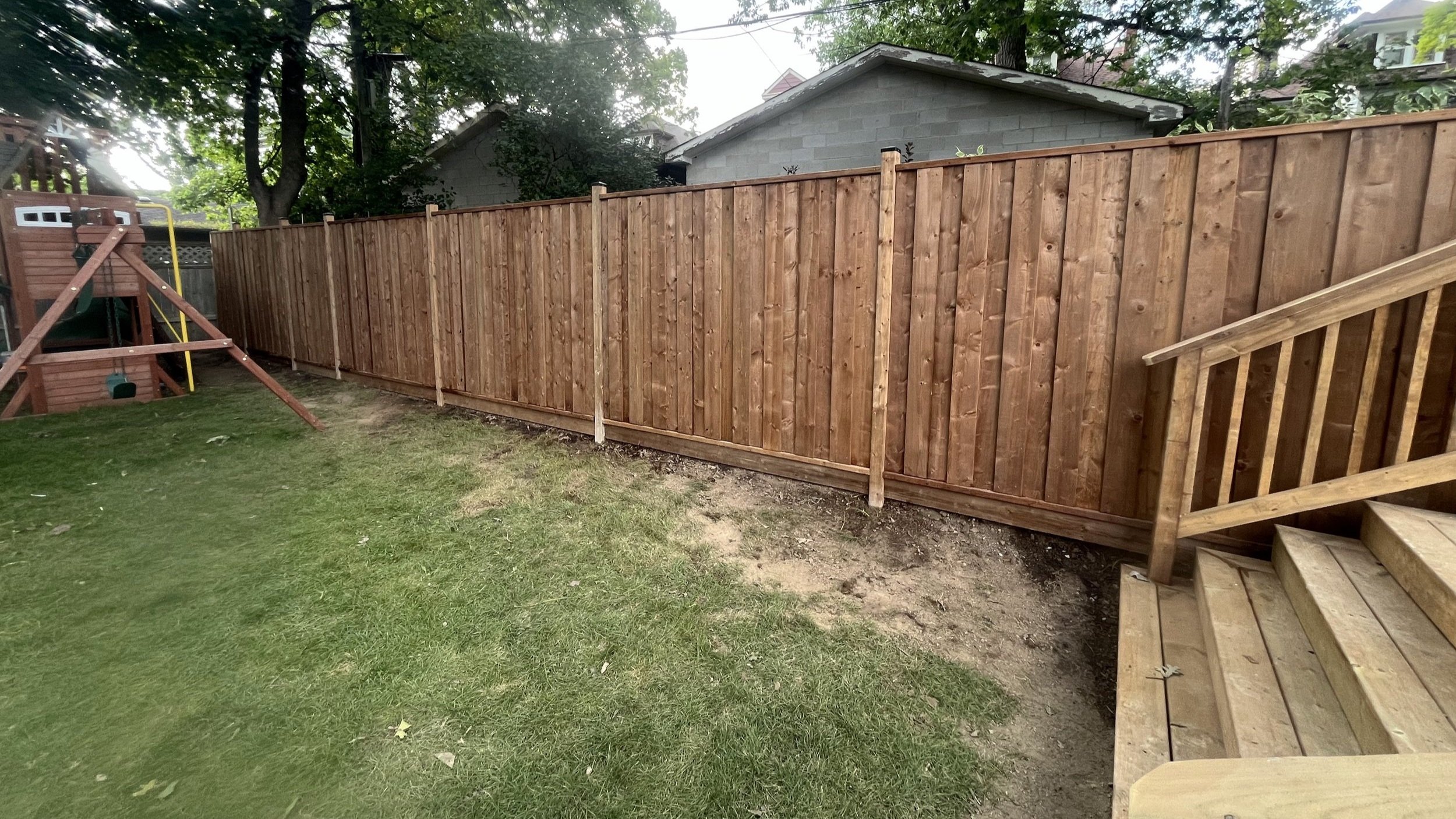 New wooden fence installed along a backyard, with a wooden staircase leading up from the grass. The yard has some patches of bare soil, and there are trees and neighboring houses in the background.