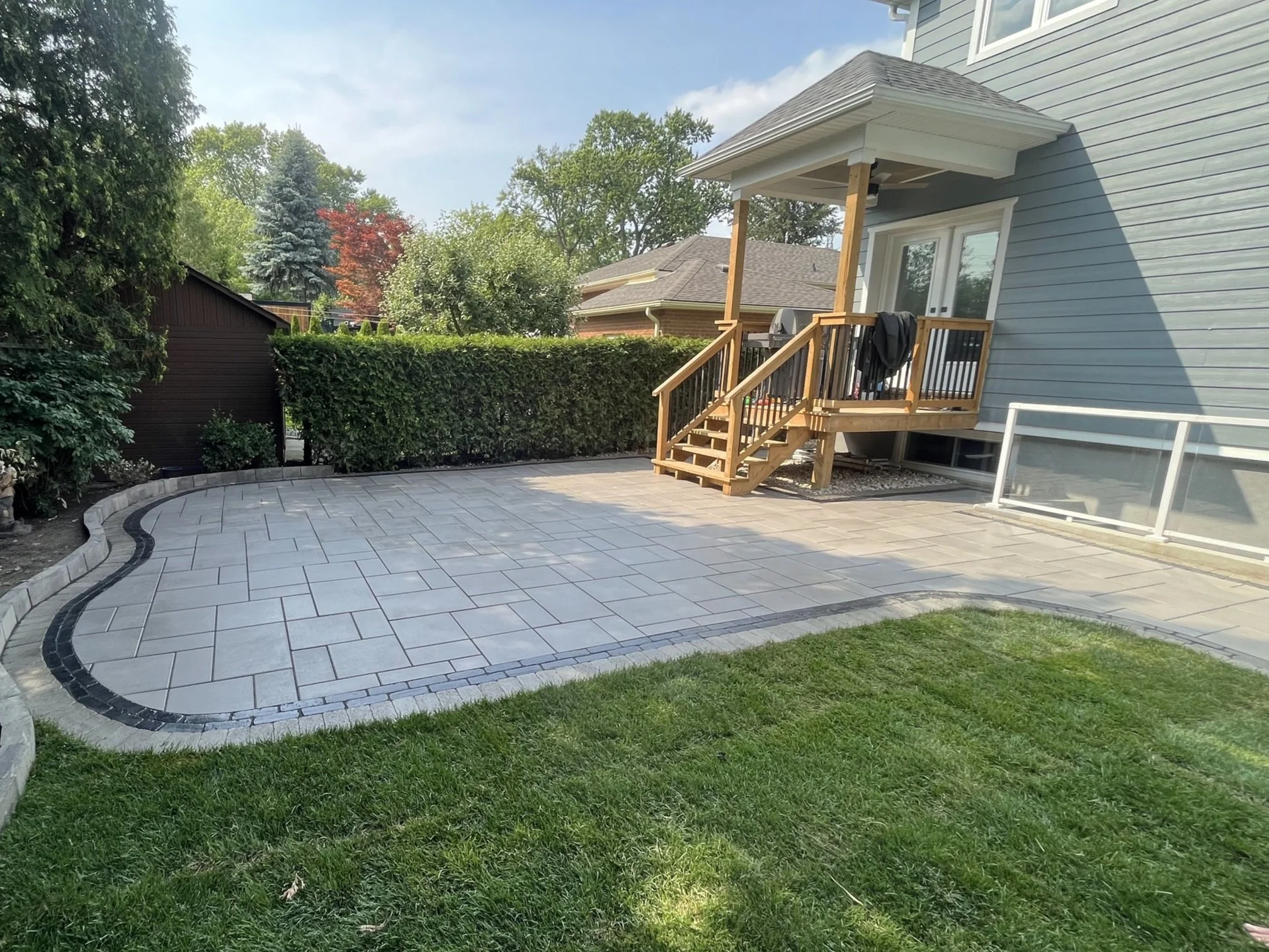 Newly built outdoor patio area with stone tiles, wooden stairs leading to a house porch, and a hedge in the background.