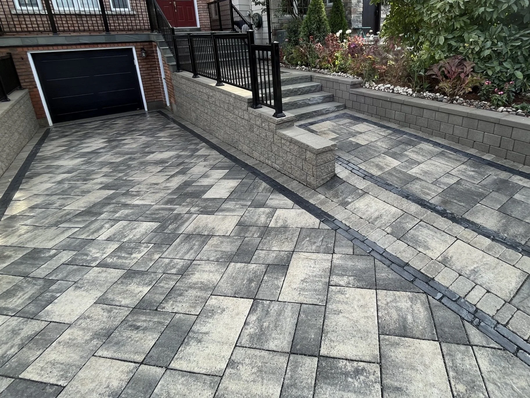 A modern residential driveway paved with interlocking stone bricks in gray tones, bordered by black stones, with a garage door and steps leading to a raised garden bed with plants and flowers.