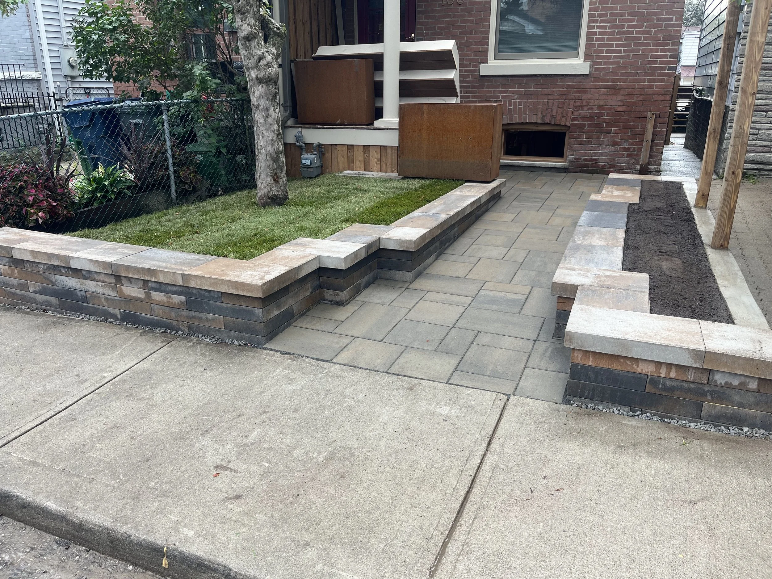Front yard with brick and stone retaining walls, new pavement, small grassy area, and garden bed with trees and plants.