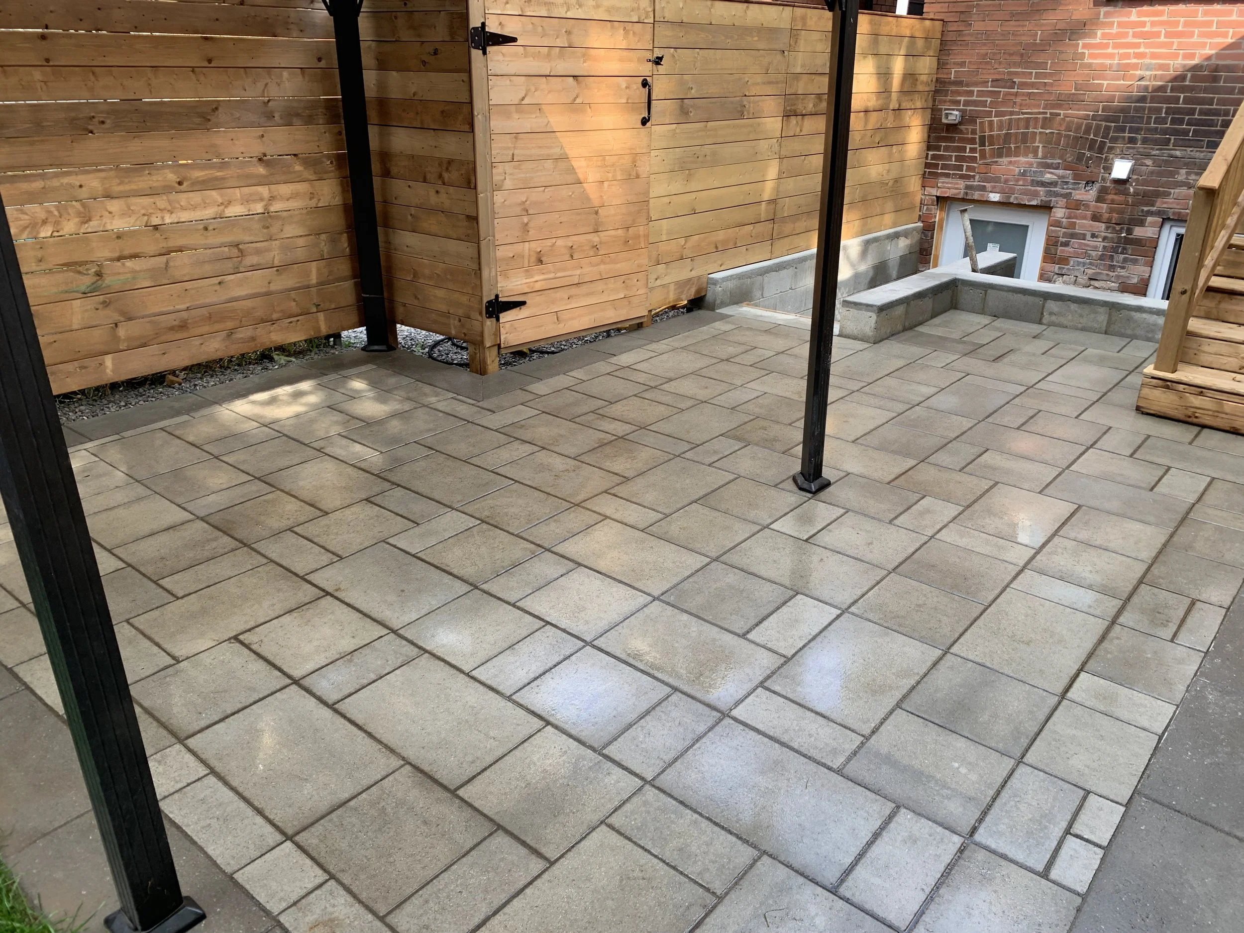 A newly constructed backyard patio with interlocking concrete pavers, surrounded by a wooden privacy fence and brick wall, with stairs leading up to the house and a small brick retaining wall.